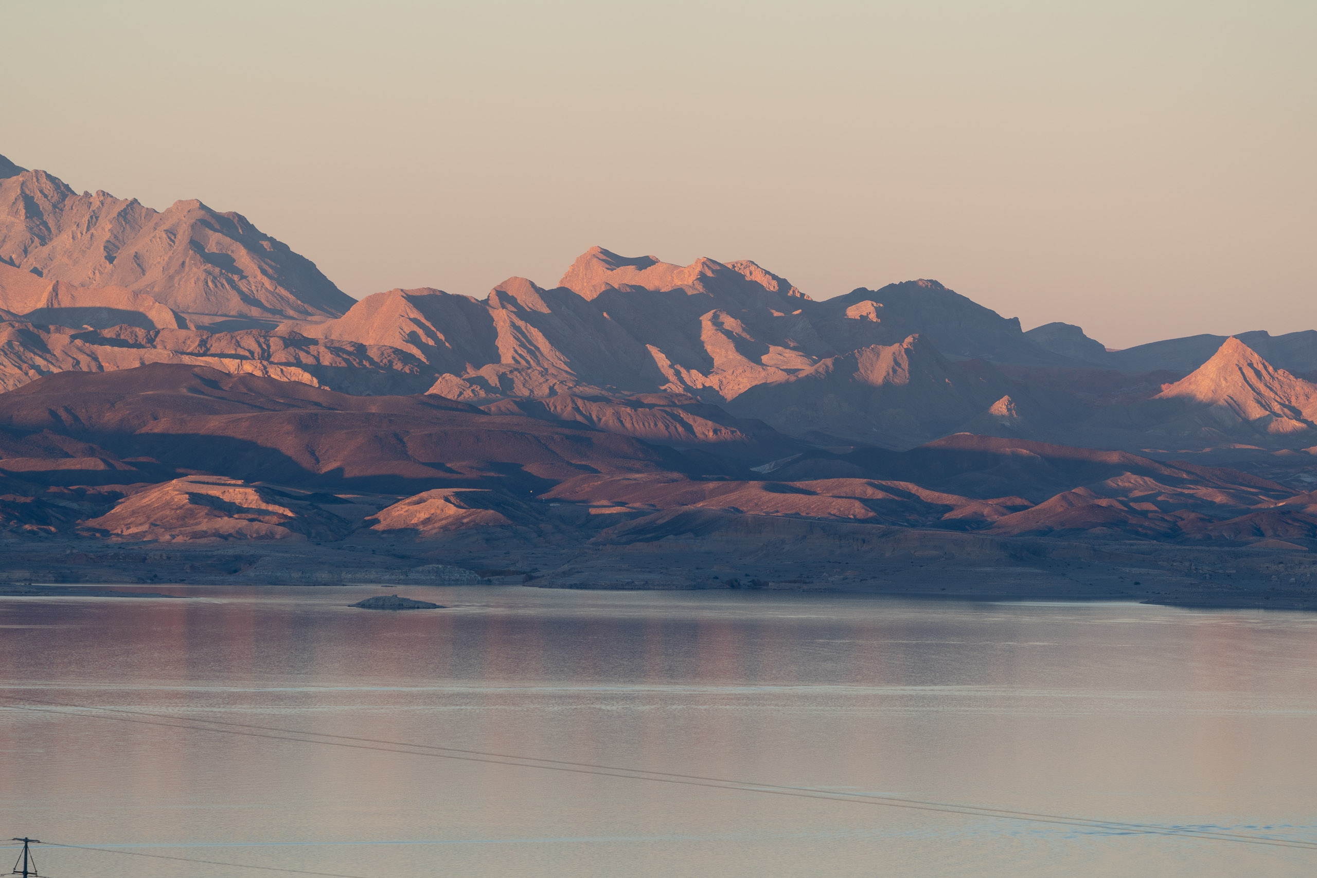 Rugged desert mountains bathed in warm sunset light rise beyond a still, reflective lake under a pale sky, with a small island visible in the middle distance