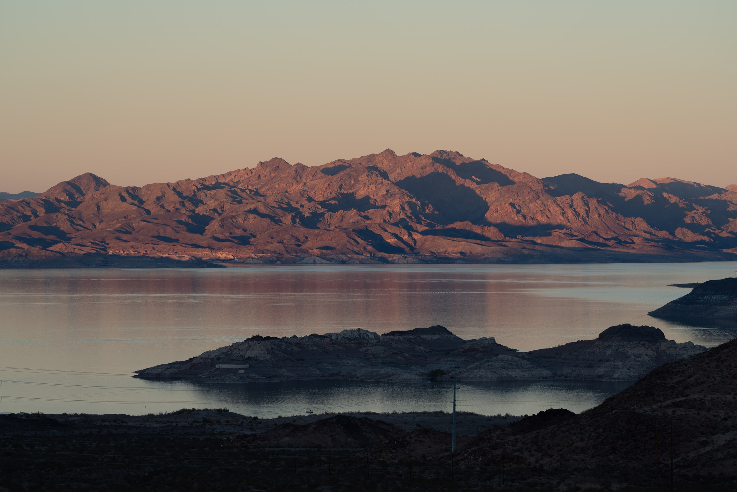 Rugged desert mountains rise above still waters at dawn, with dark rocky islands dotting the pale lake under a soft, muted sky