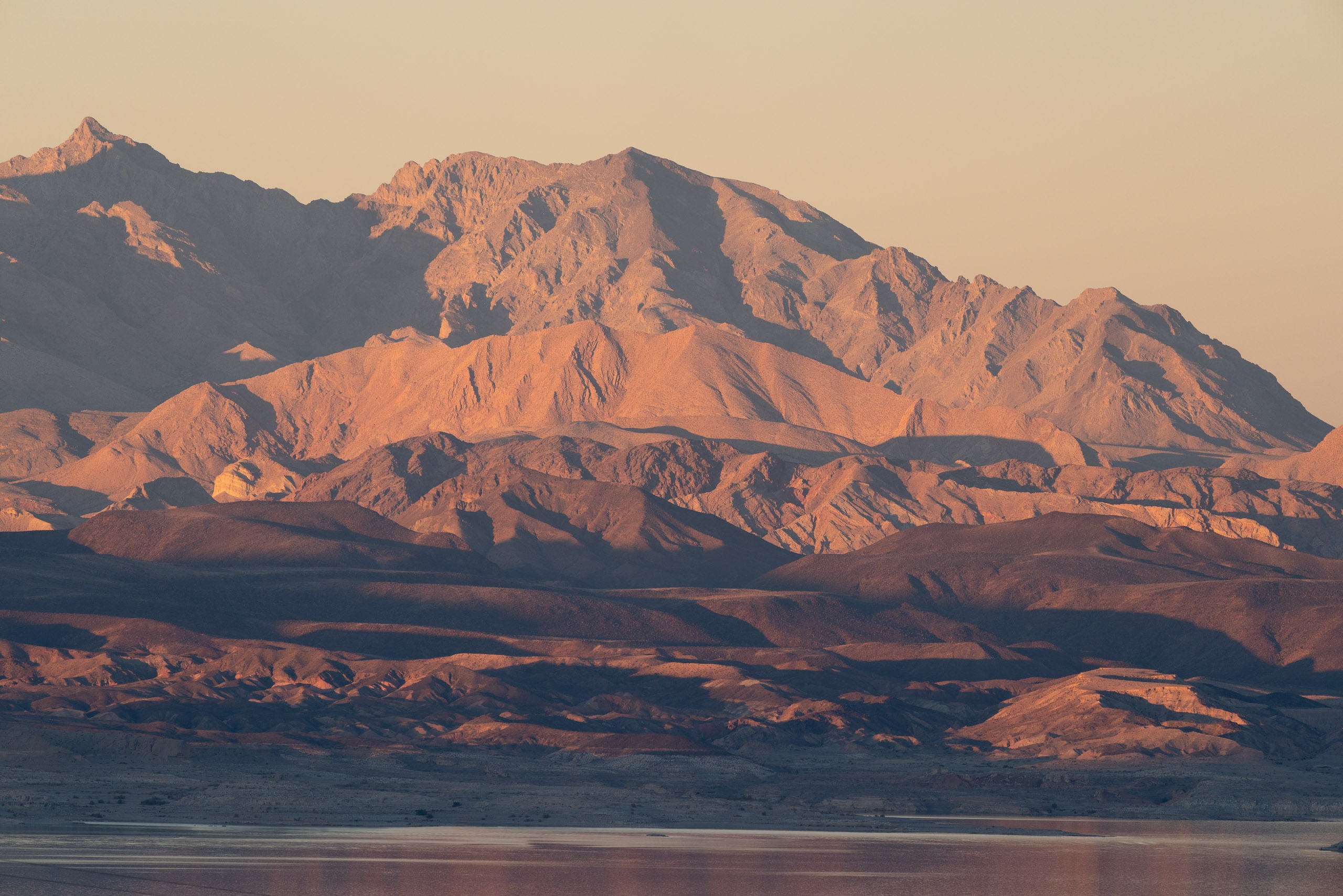 Golden sunlight illuminates rugged desert mountains above a calm lake, with layered ridges casting deep shadows across the arid landscape at dawn or dusk