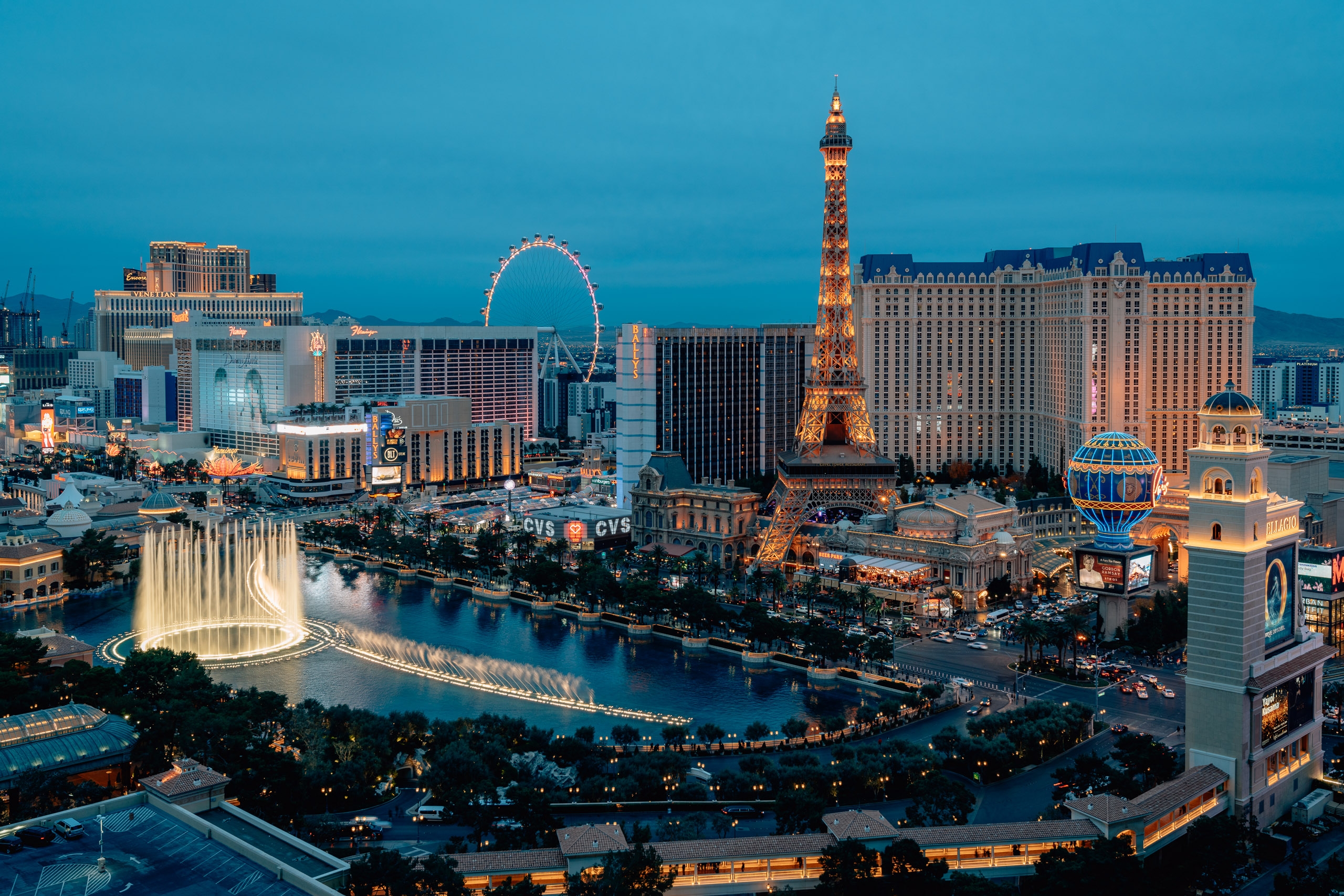 Las Vegas skyline at dusk features the illuminated Eiffel Tower replica, Bellagio fountains, and High Roller observation wheel against a deep blue twilight sky