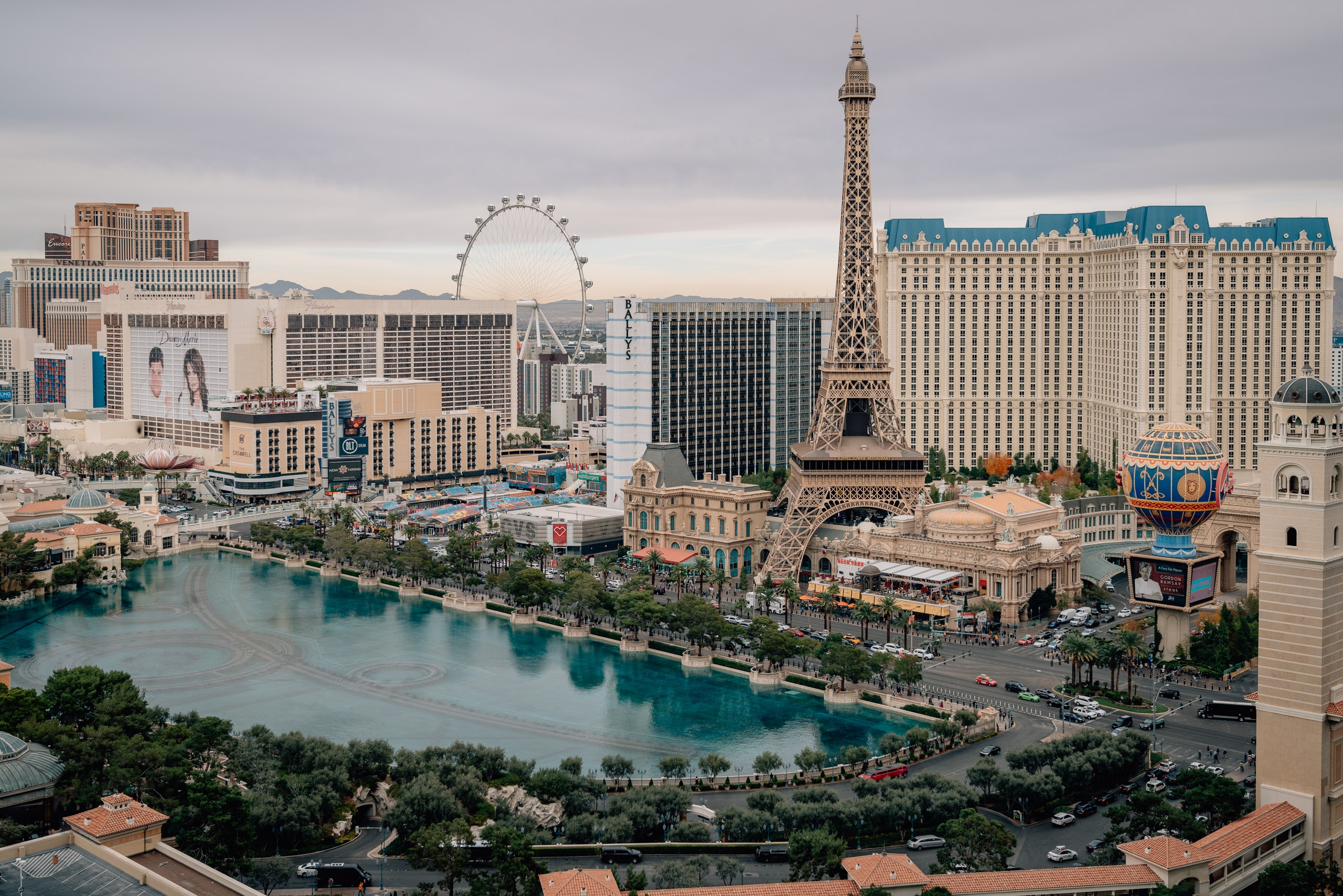 The Las Vegas Strip stretches across the frame featuring the Eiffel Tower replica, Bellagio fountain with turquoise water, the Ferris wheel, and surrounding hotels under an overcast sky