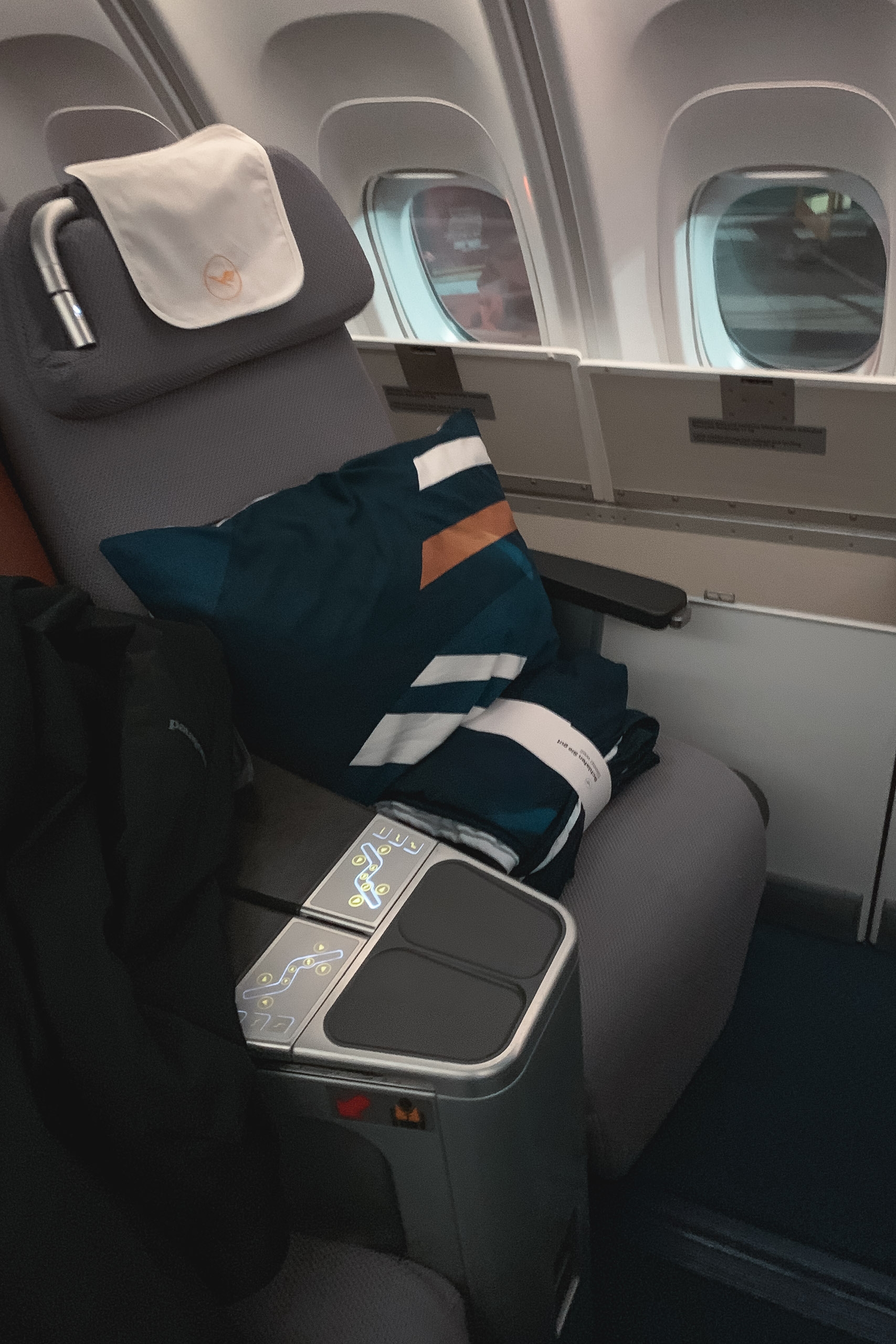 An airline seat holds a folded navy blanket with white and orange stripes, with a small pillow and headphones on the headrest, windows visible behind in daylight