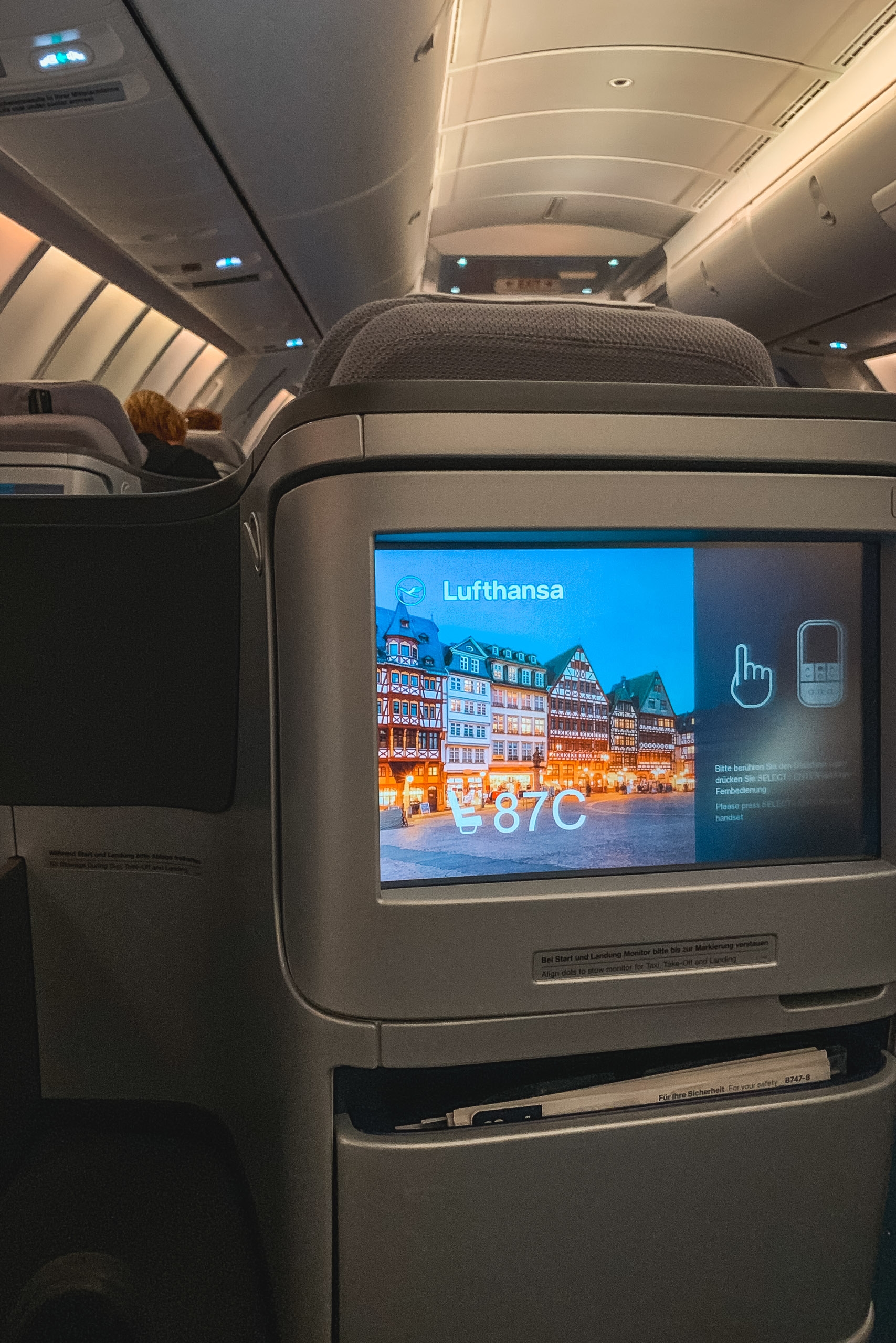 Airplane cabin interior with a seatback entertainment screen displaying a Lufthansa advertisement featuring Frankfurt's illuminated historic district and temperature information, with overhead bins and cabin ceiling visible above