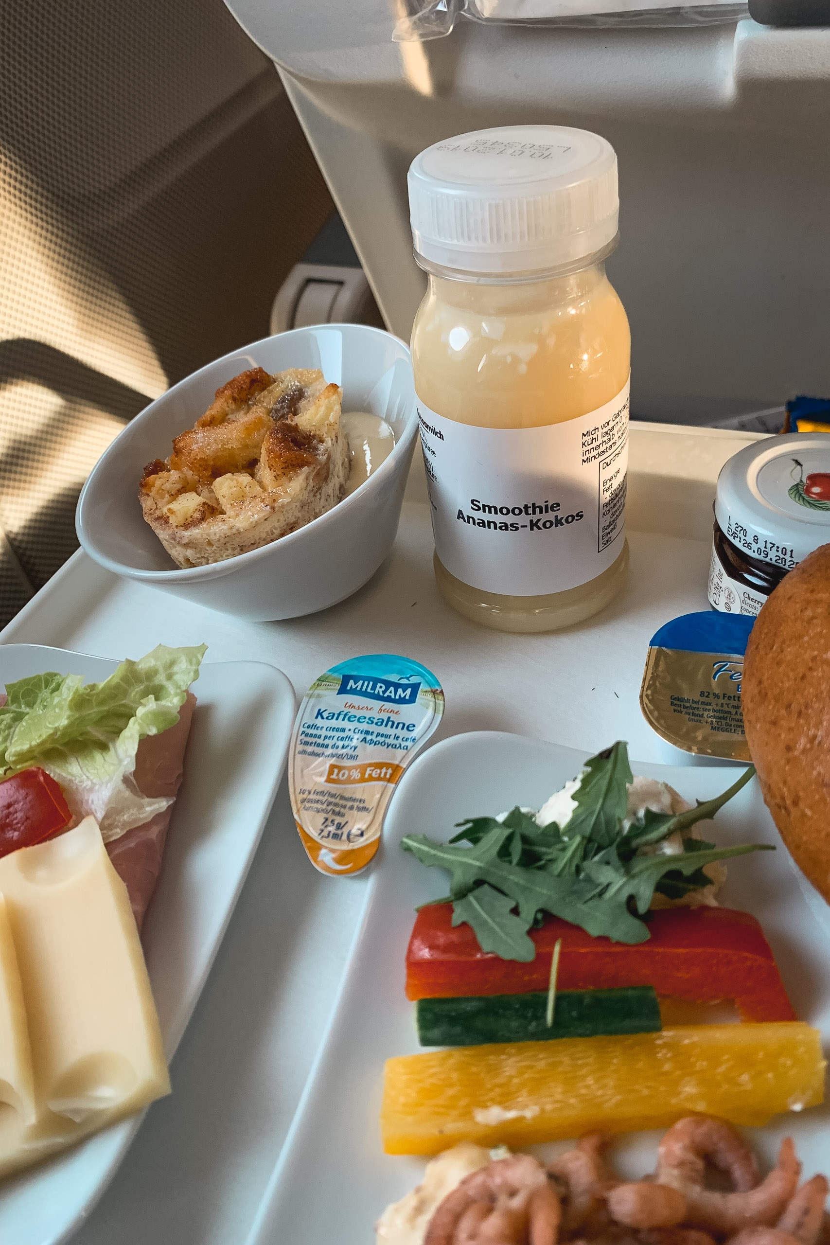 An airplane breakfast tray displays fresh pastries, cheese, vegetables, pineapple-coconut smoothie, and butter portions in bright natural window light during flight