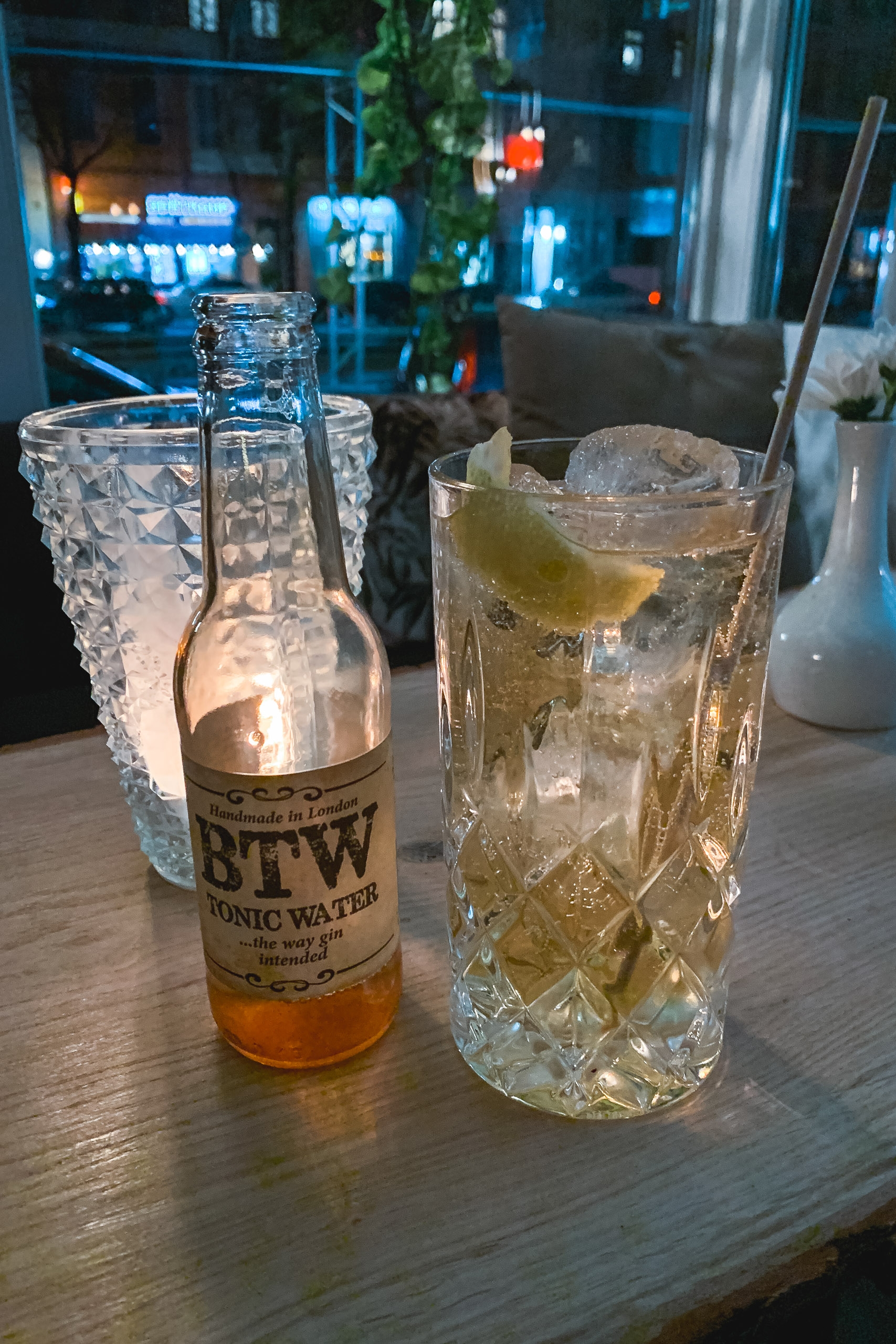 A bottle of BTW tonic water sits beside a tall glass filled with amber liquid and ice, lemon slice visible, set on a wooden bar counter with blurred city lights and neon signs glowing through the window behind