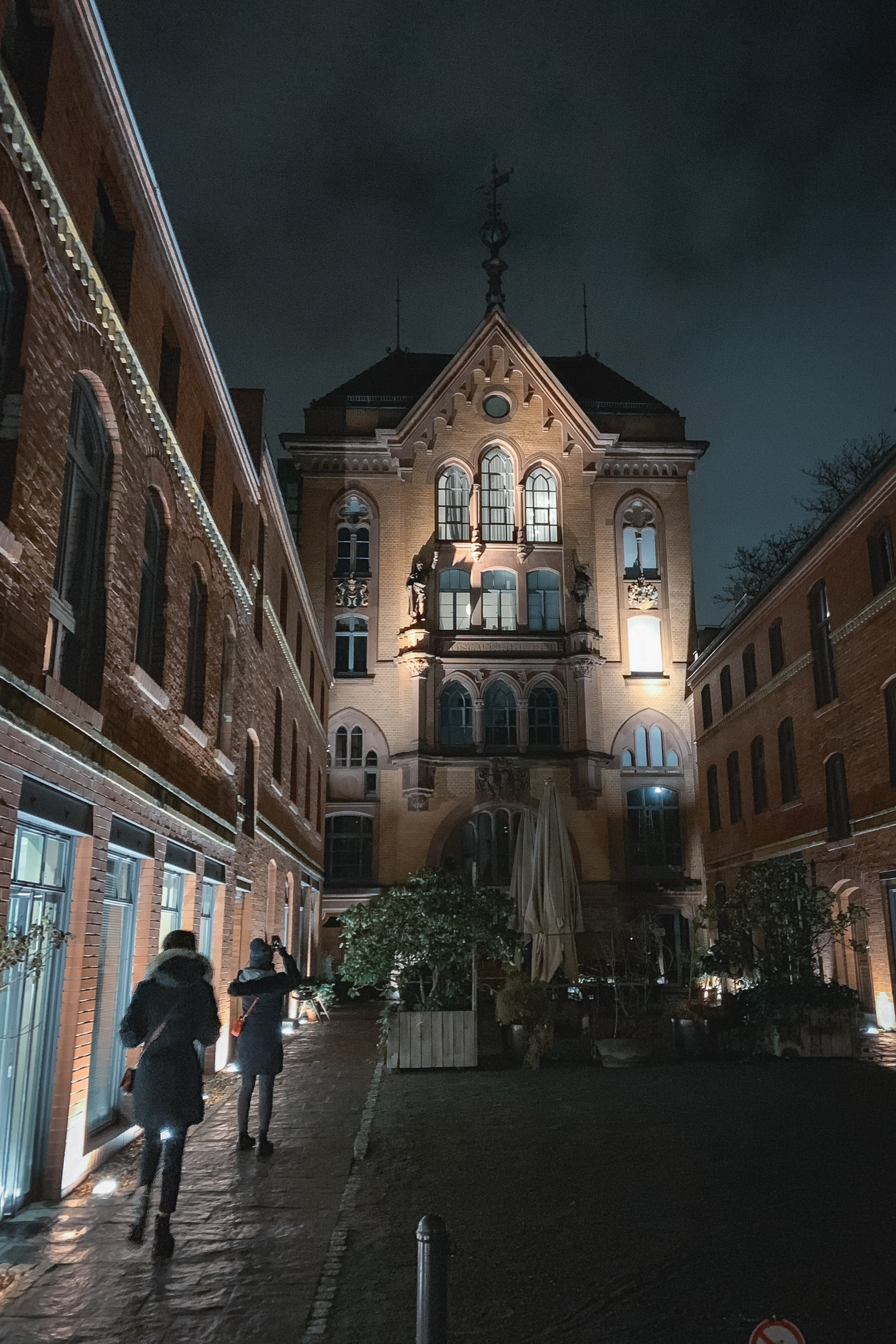 A narrow European courtyard at dusk features a illuminated brick tower with ornate architecture, flanked by residential buildings, while two figures walk on the wet cobblestone street below a dramatic stormy sky