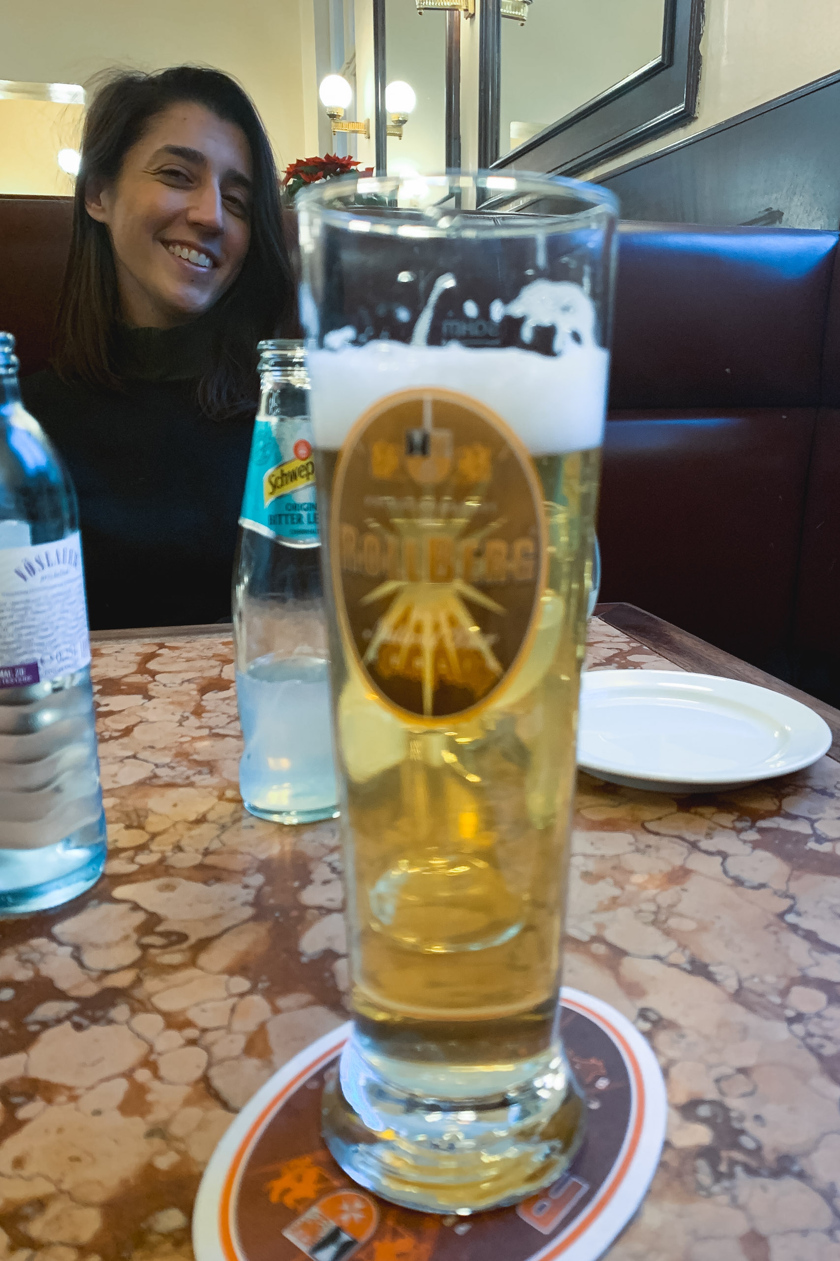 A woman smiles at a restaurant table where a tall glass of pale beer with a foamy head sits in the foreground, with bottles and place settings visible in the dimly lit interior space