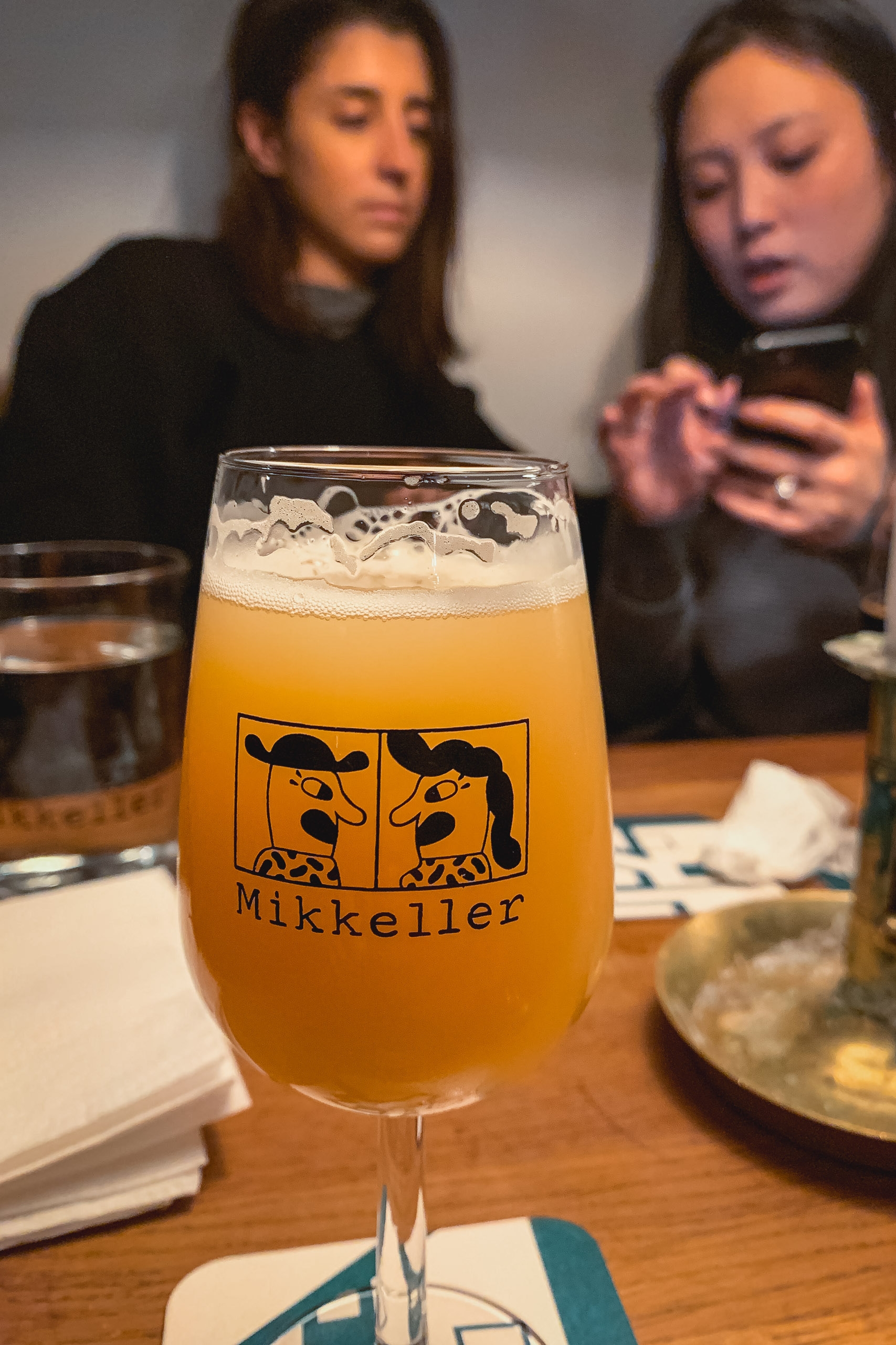 A Mikkeller beer glass filled with golden ale sits in sharp focus on a wooden table while two women blur in the background, one checking her phone in warm interior lighting