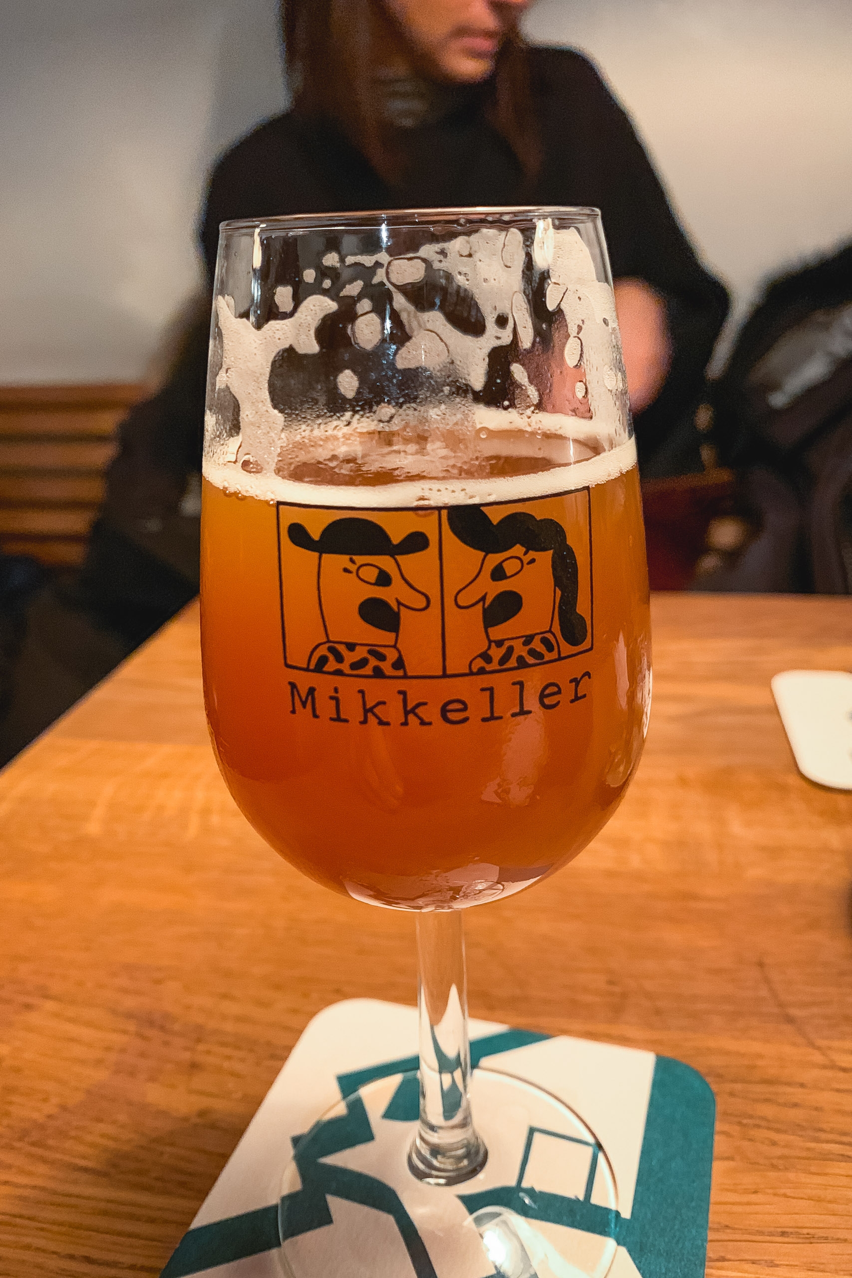 A Mikkeller brewery glass filled with amber beer and foam sits on a wooden table, with a blurred person in dark clothing visible in the background