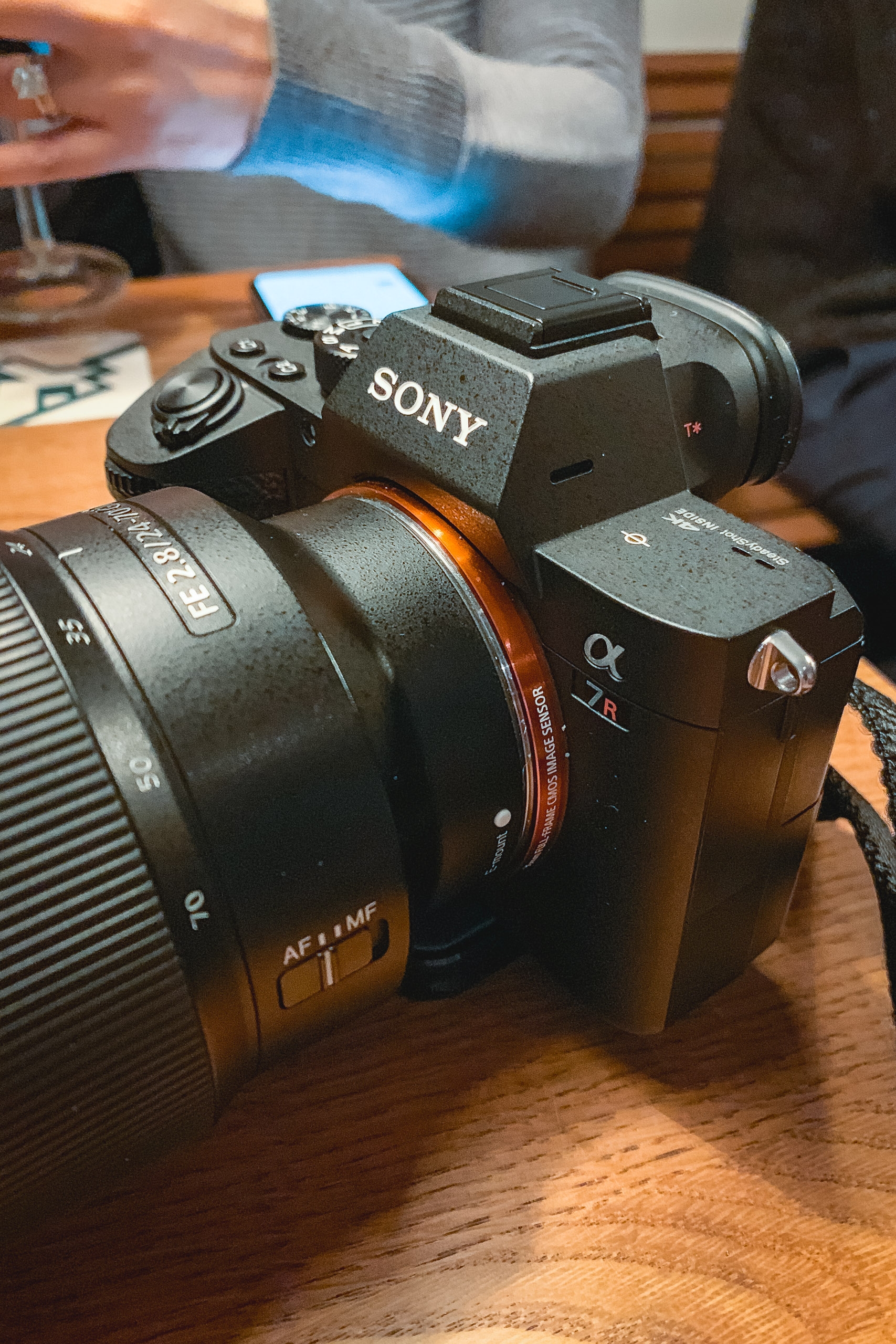A Sony mirrorless camera with attached lens sits on a wooden table indoors, with a blurred hand adjusting equipment in the warm background lighting