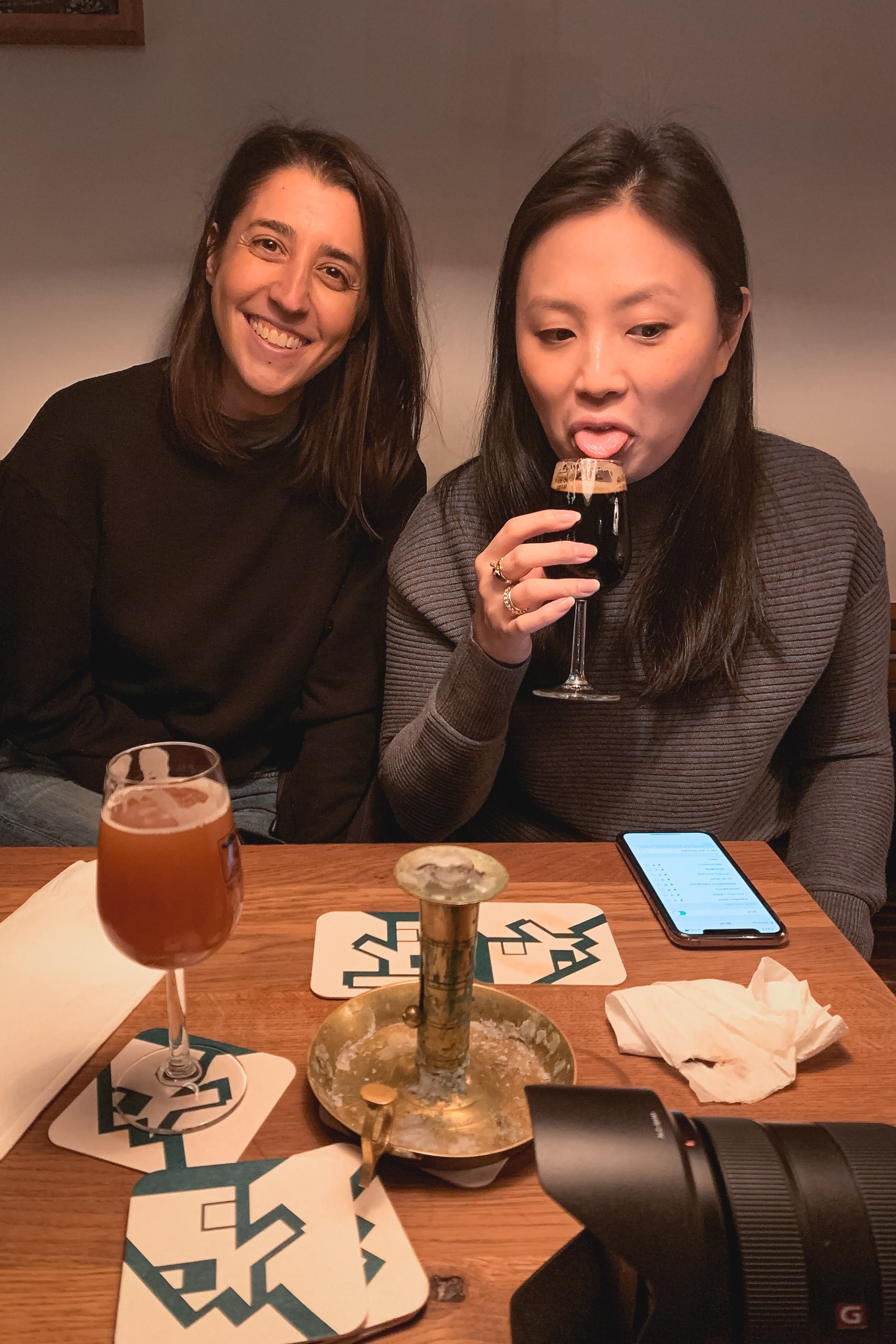 Two women sit at a wooden table, one smiling at the camera while the other drinks from a glass, surrounded by cocktails, a smartphone, and napkins in warm indoor lighting