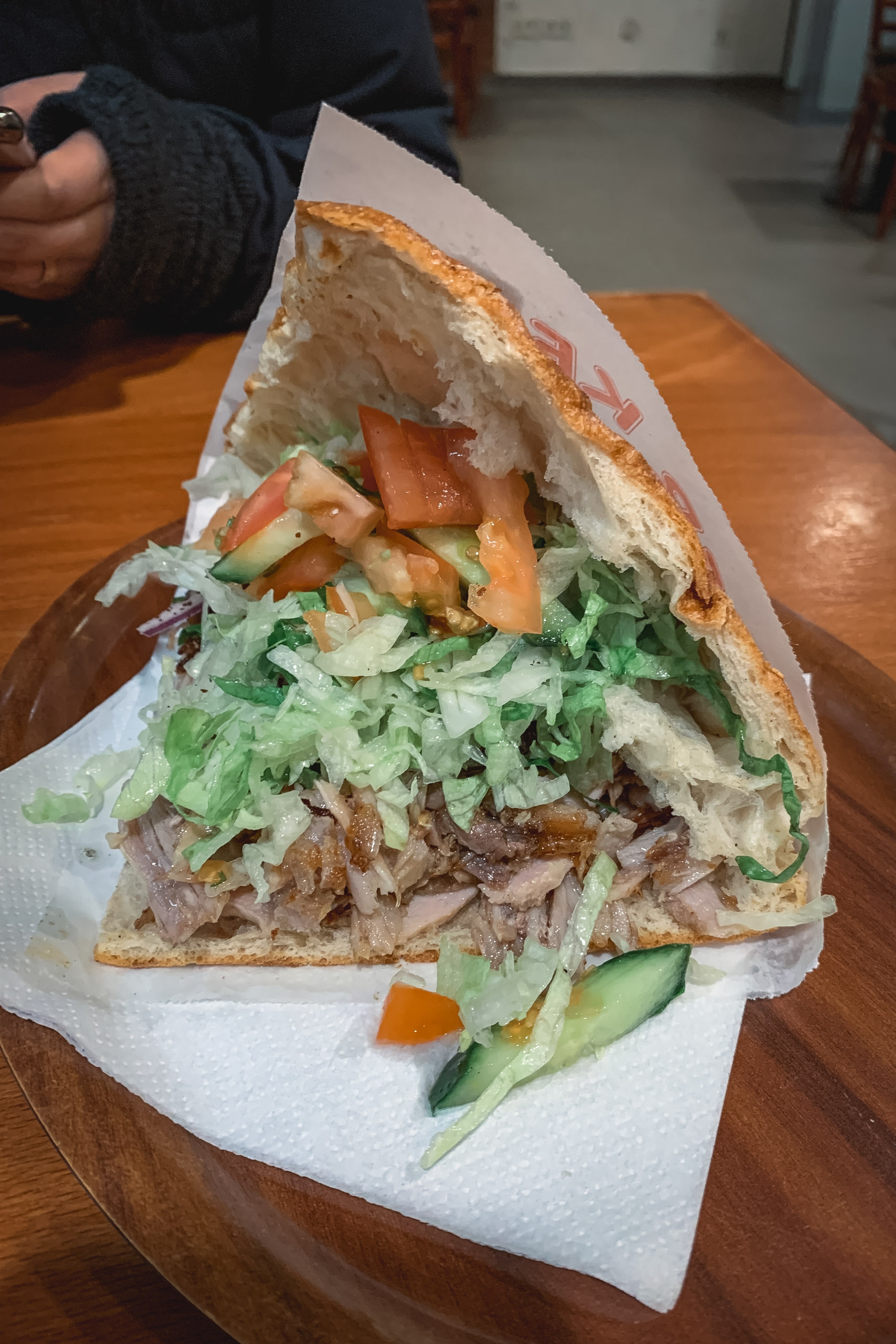 A person's hand holds a crusty sandwich wrapped in white paper, filled with sliced meat, fresh greens, tomato, and cucumber, sitting on a wooden table in an indoor setting