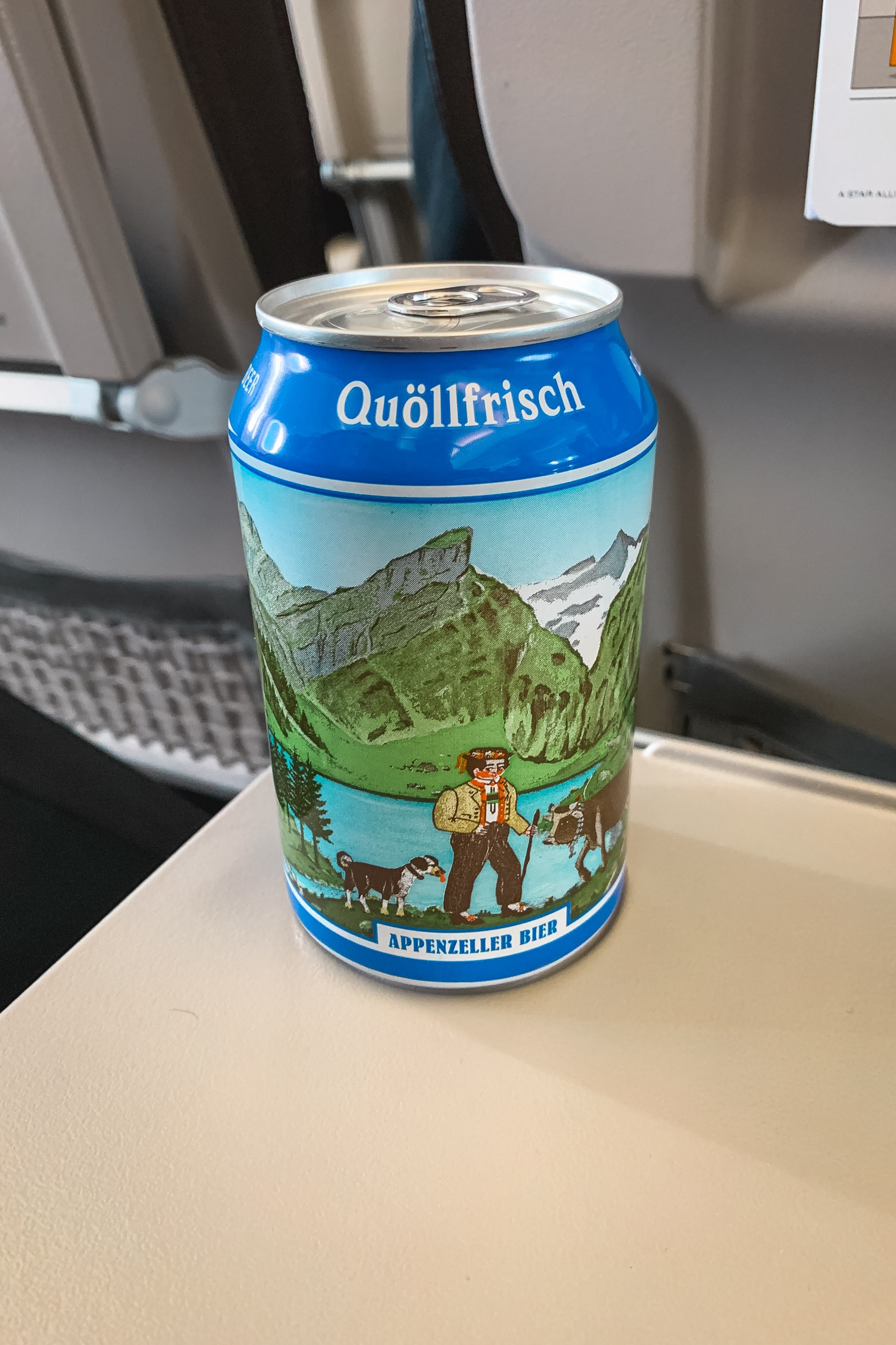 A Quöllfrisch Appenzeller beer can sits on a white desk in an office setting, featuring Alpine mountain scenery and traditional Swiss imagery on its label