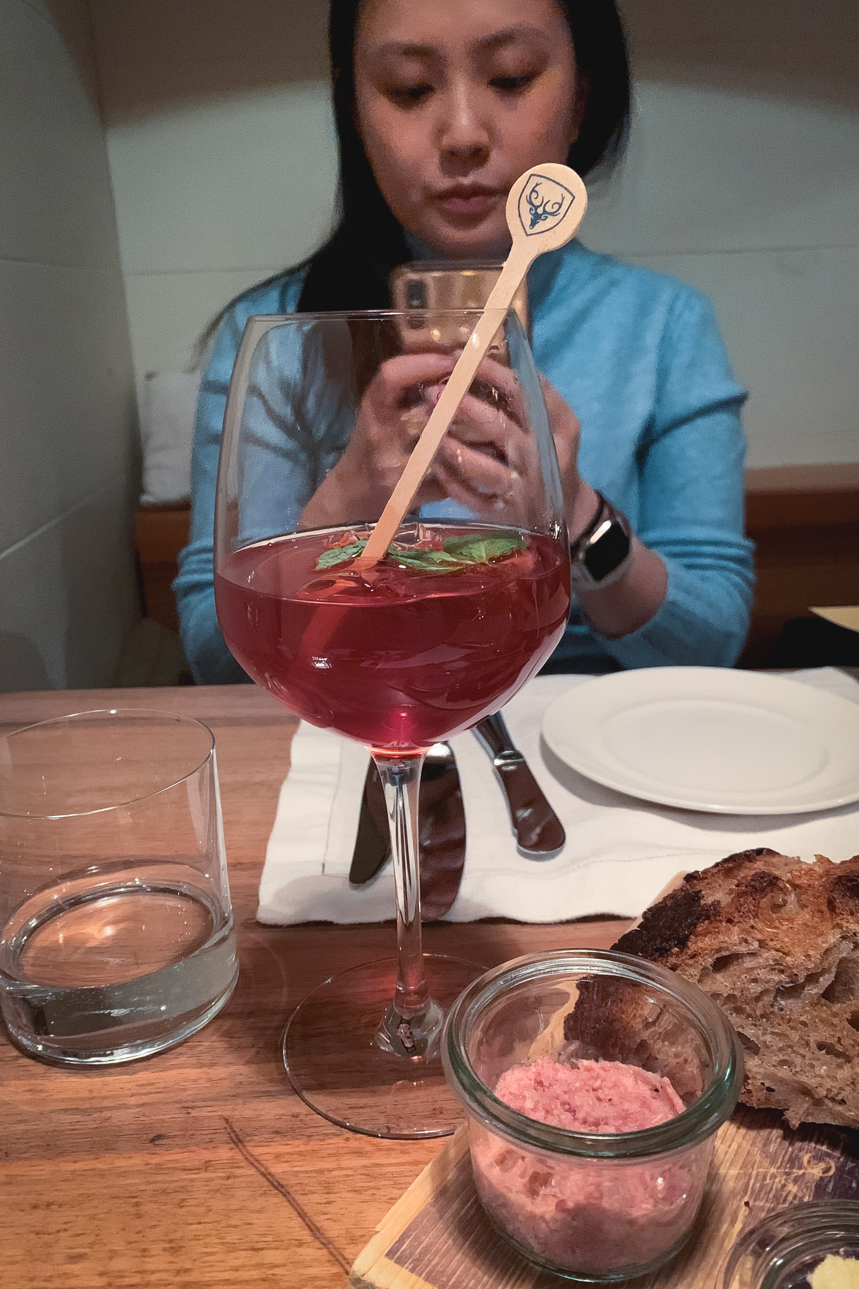 A woman in a blue shirt stirs a deep red cocktail with a wooden spoon at a dining table, with bread and small dishes visible beside her in warm indoor lighting
