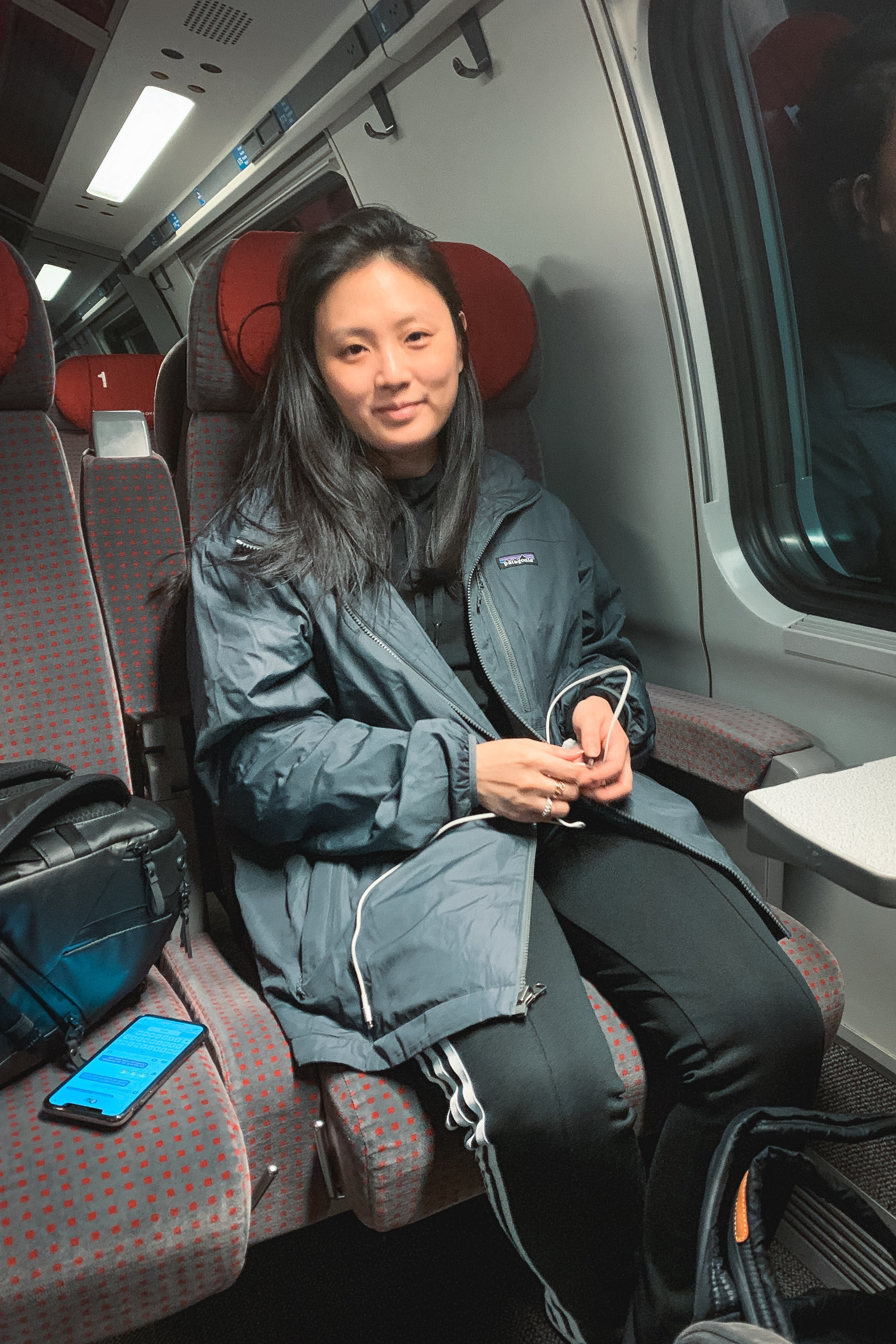 A young woman sits in a red-upholstered train seat wearing a blue jacket, holding white earbuds, with a smartphone visible beside her in the modern transit car interior