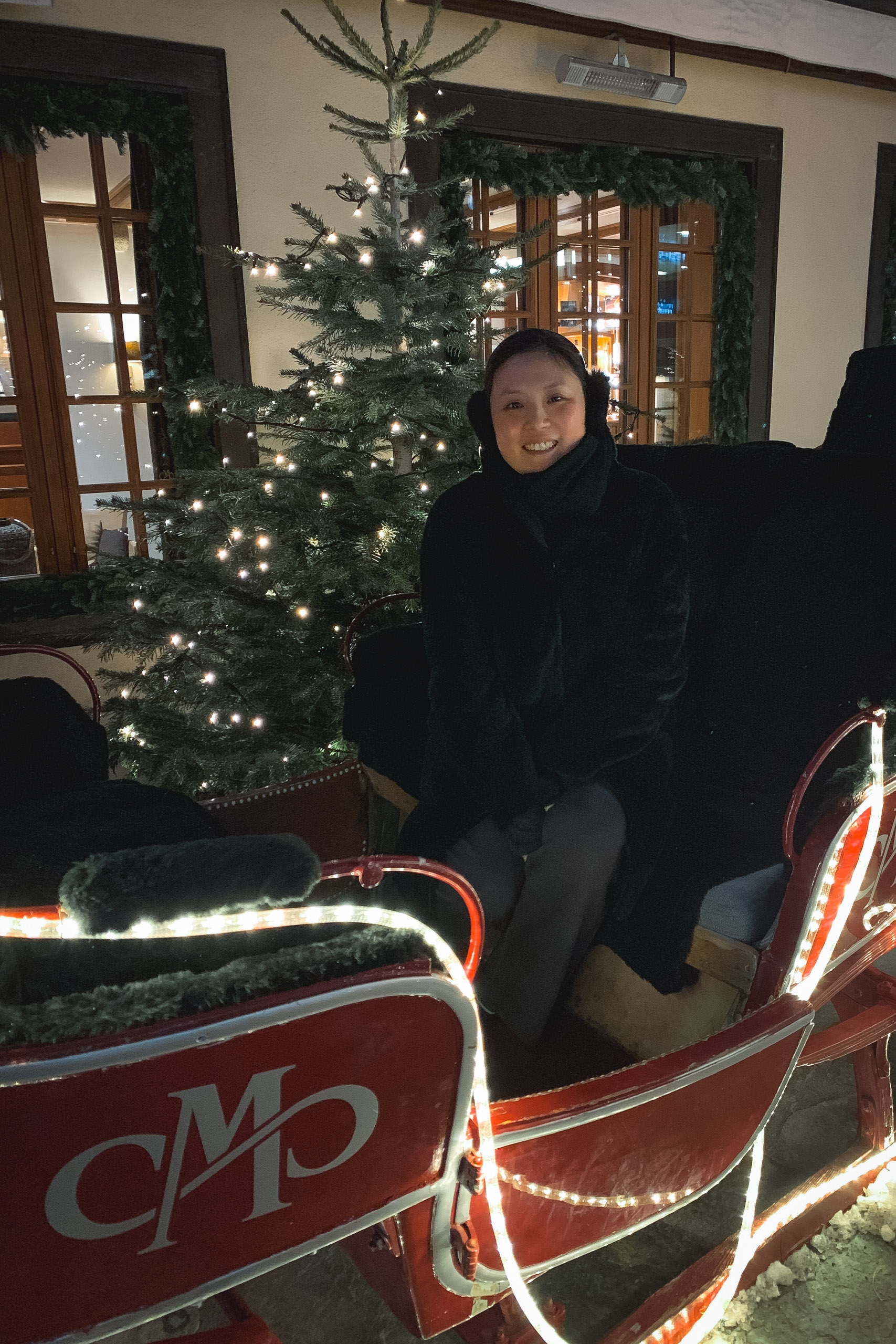 Woman in black winter clothing poses beside an illuminated decorative sleigh with glowing rope lights in front of a Christmas tree indoors.