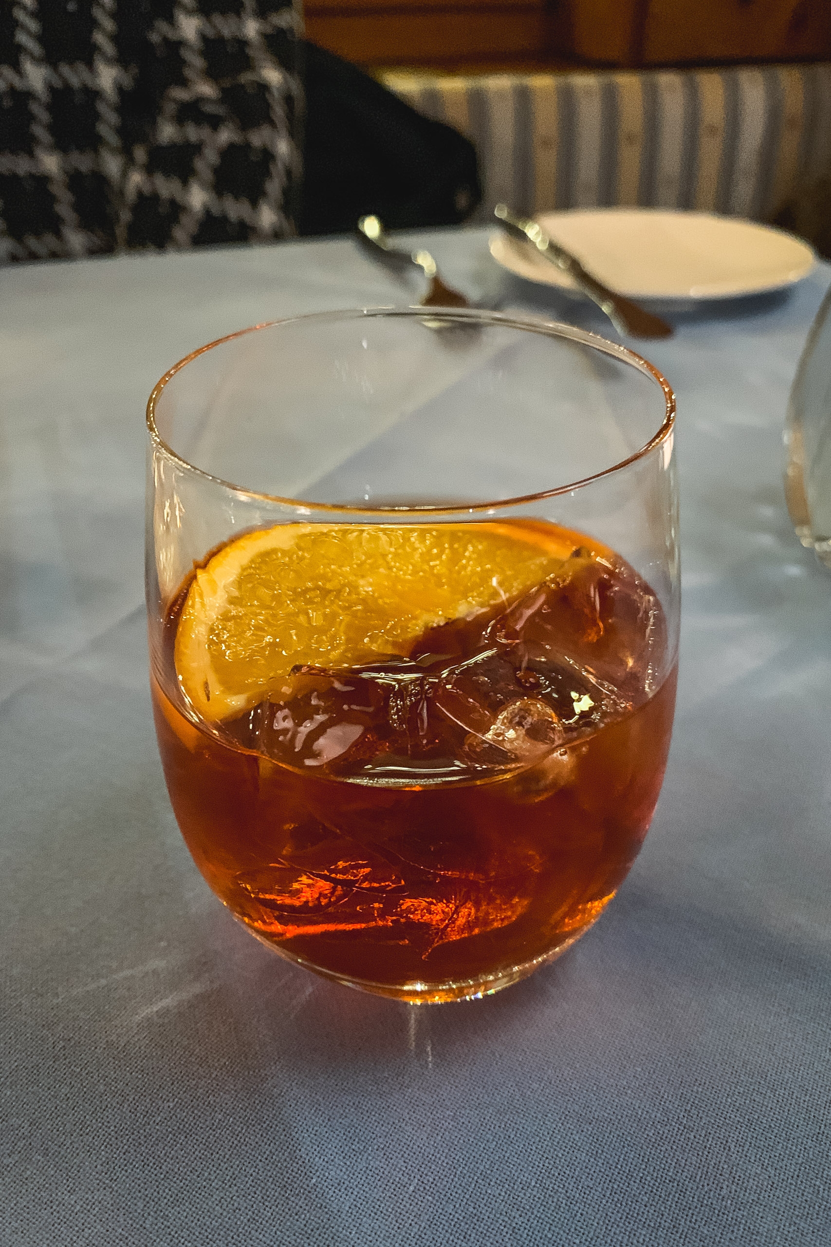 A cocktail with amber liquid and ice is garnished with an orange slice, sitting on a white tablecloth in an upscale dining setting with blurred diners and striped seating in the background