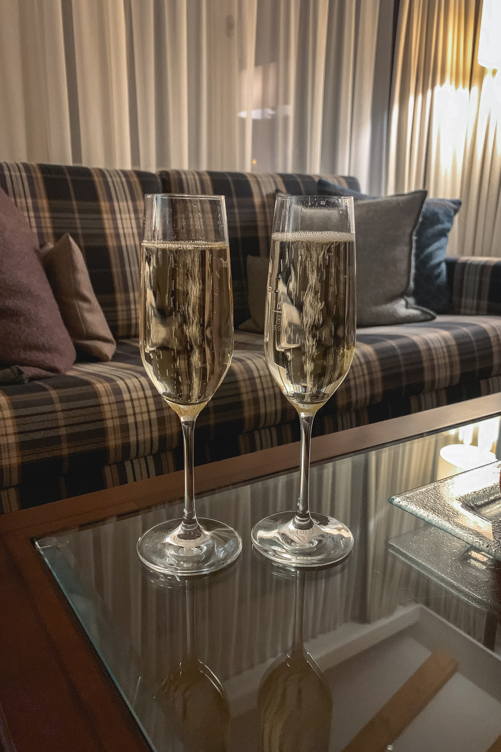 Two champagne flutes filled with sparkling wine sit on a glass table in front of a plaid sofa, with warm sunlight streaming through sheer curtains