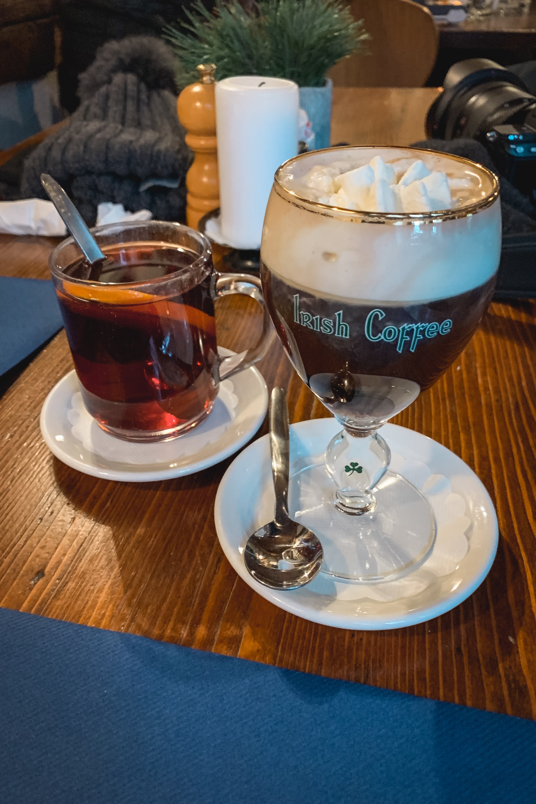 Two Irish coffee drinks sit on a wooden table with white ceramic saucers, one featuring dark espresso topped with cream foam and the other a darker brewed coffee, accompanied by potted plants and home décor in soft interior lighting.