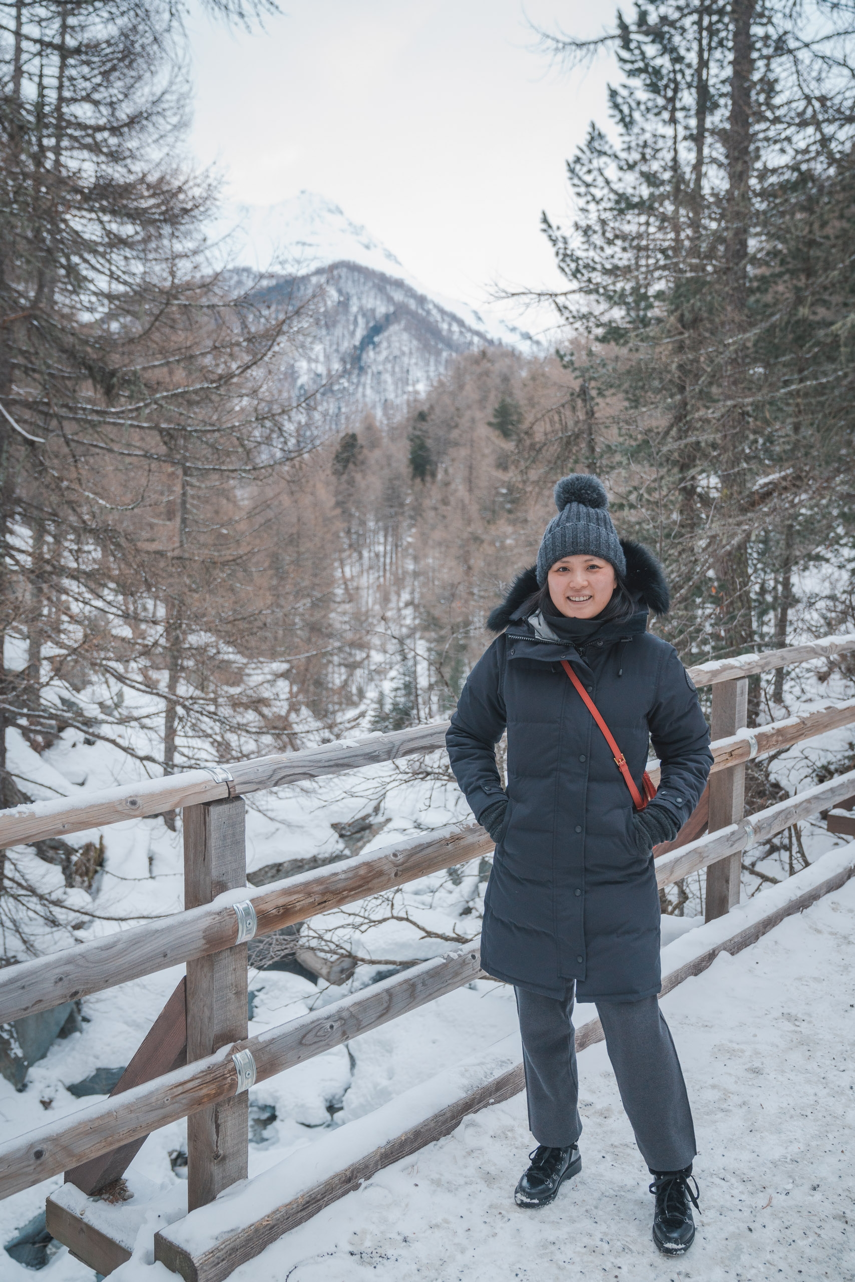 A woman in winter gear stands on a snowy wooden bridge, smiling at the camera with snow-covered alpine mountains and evergreen forests framing the scenic valley backdrop