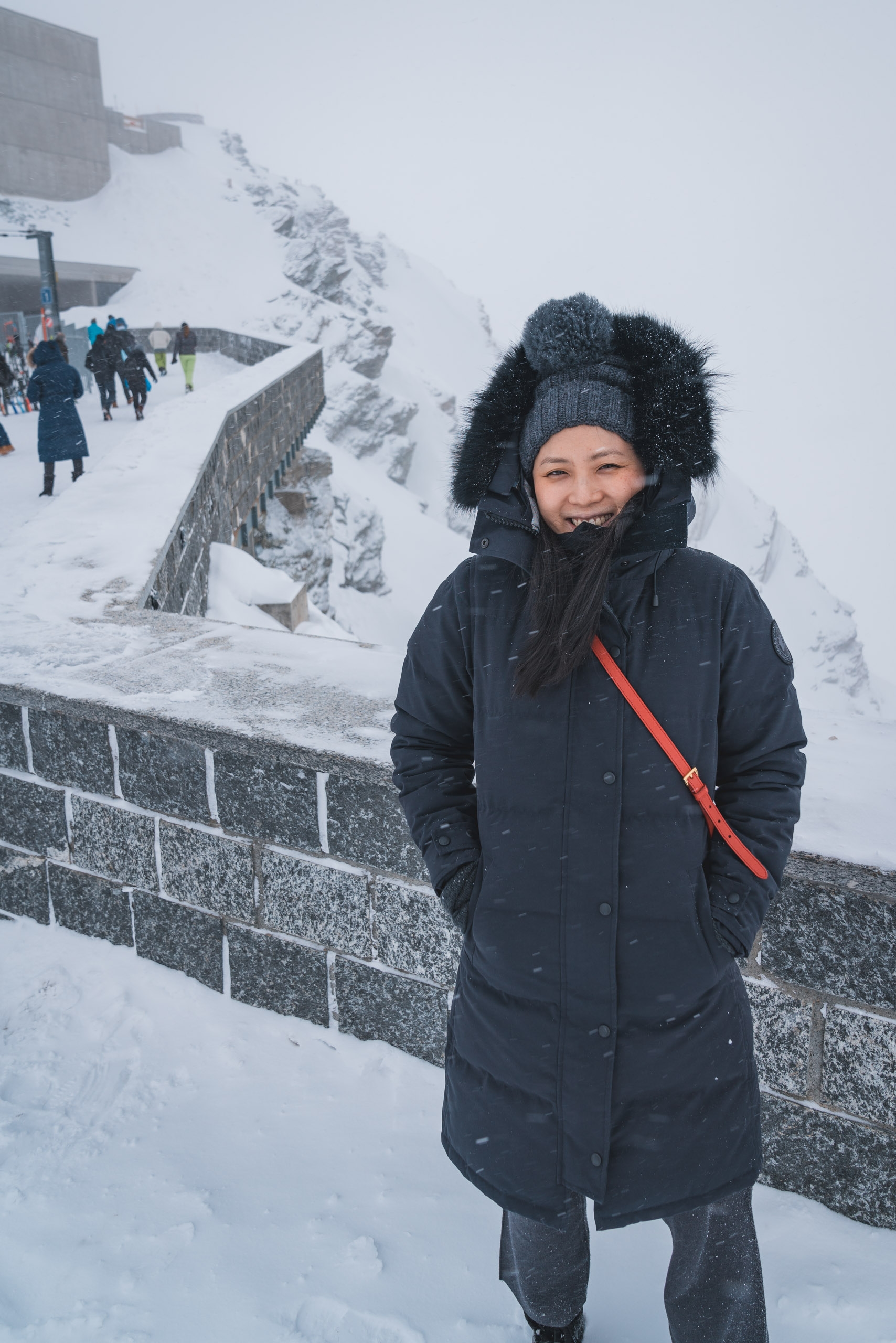 Woman in black winter coat and fuzzy ear warmer smiles at snow-covered mountain observation deck with visitors and rocky peaks in overcast background