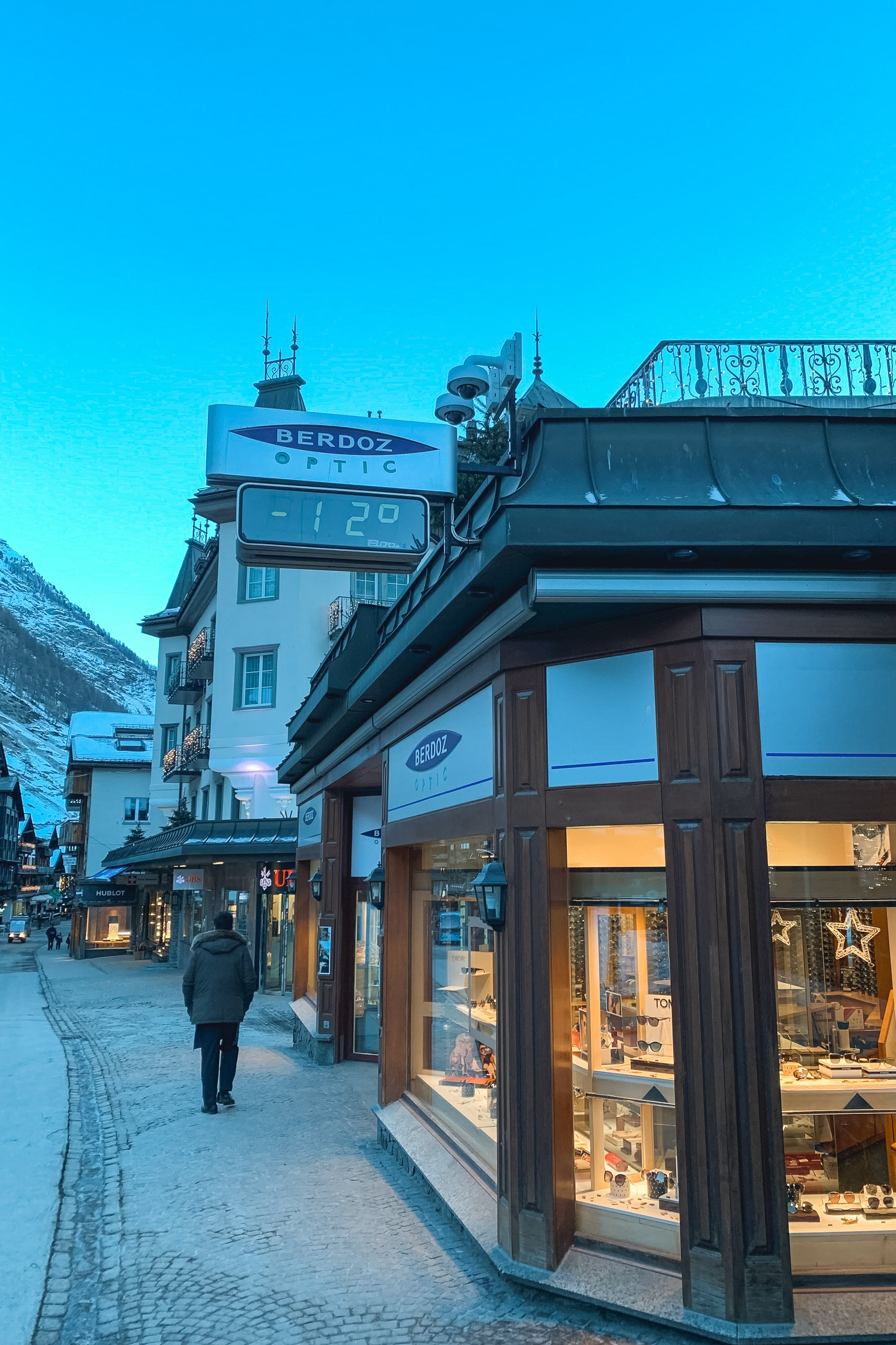Alpine town street at dusk with illuminated shop windows, a Berdoz clock tower, and pedestrians walking past traditional chalets under clear blue sky