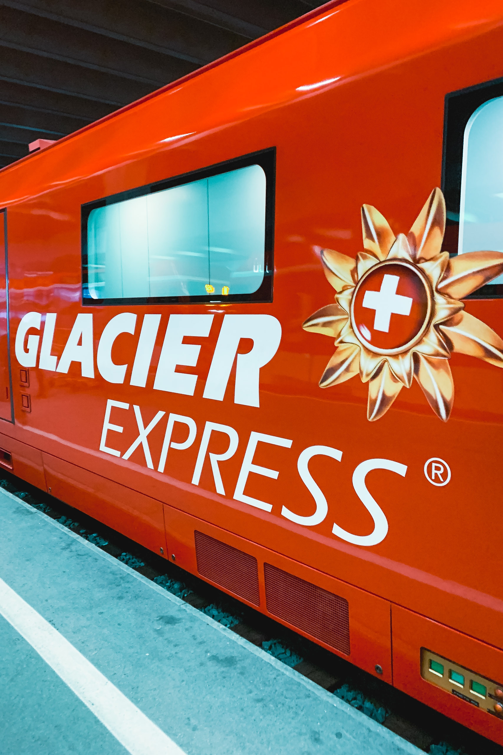 A bright red Glacier Express train car displays the company logo and Swiss cross emblem against a turquoise platform under industrial overhead structure