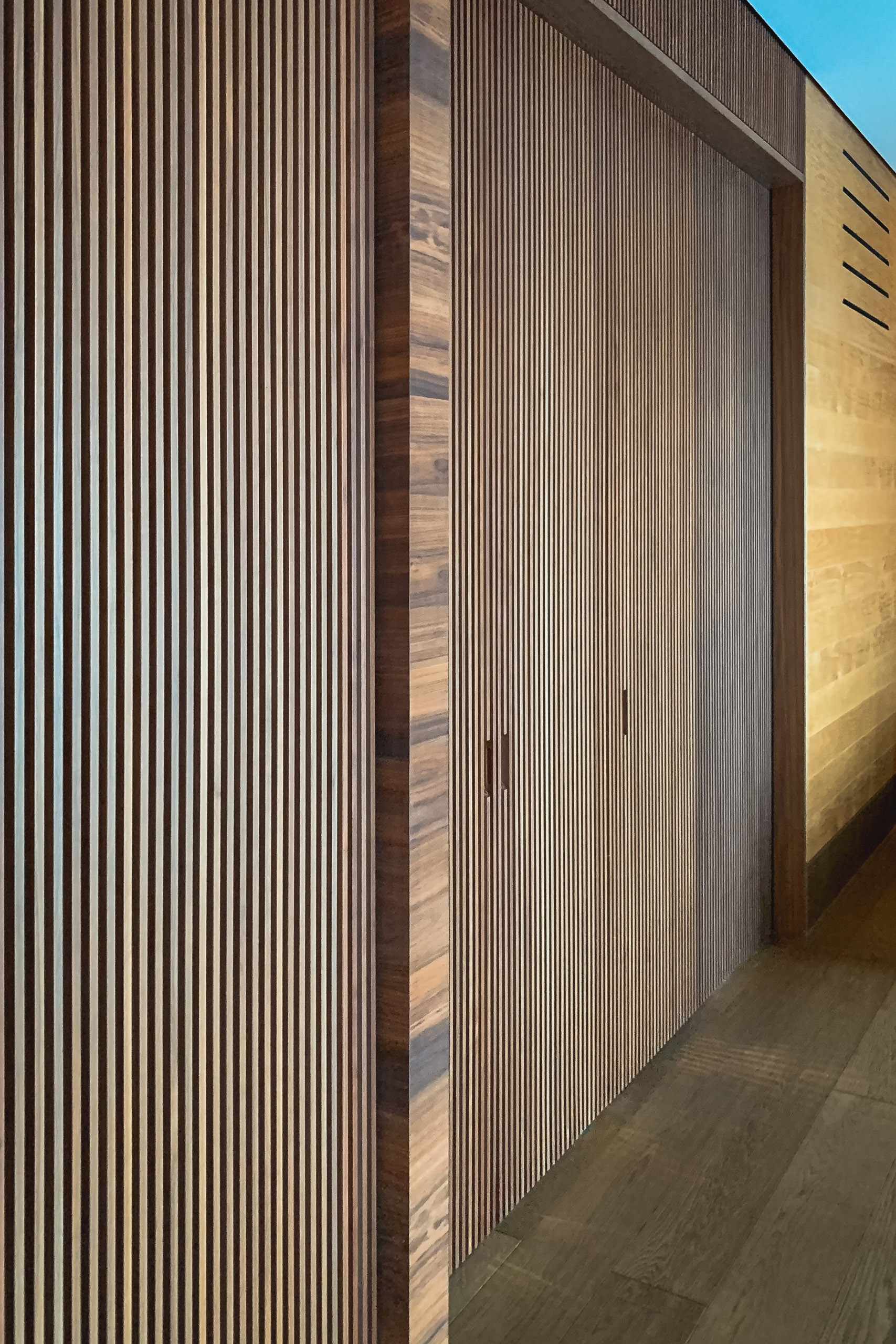 Modern architectural facade with vertical wooden slats creates strong linear patterns, accented by warm golden lighting against a clear blue sky and dark pavement below
