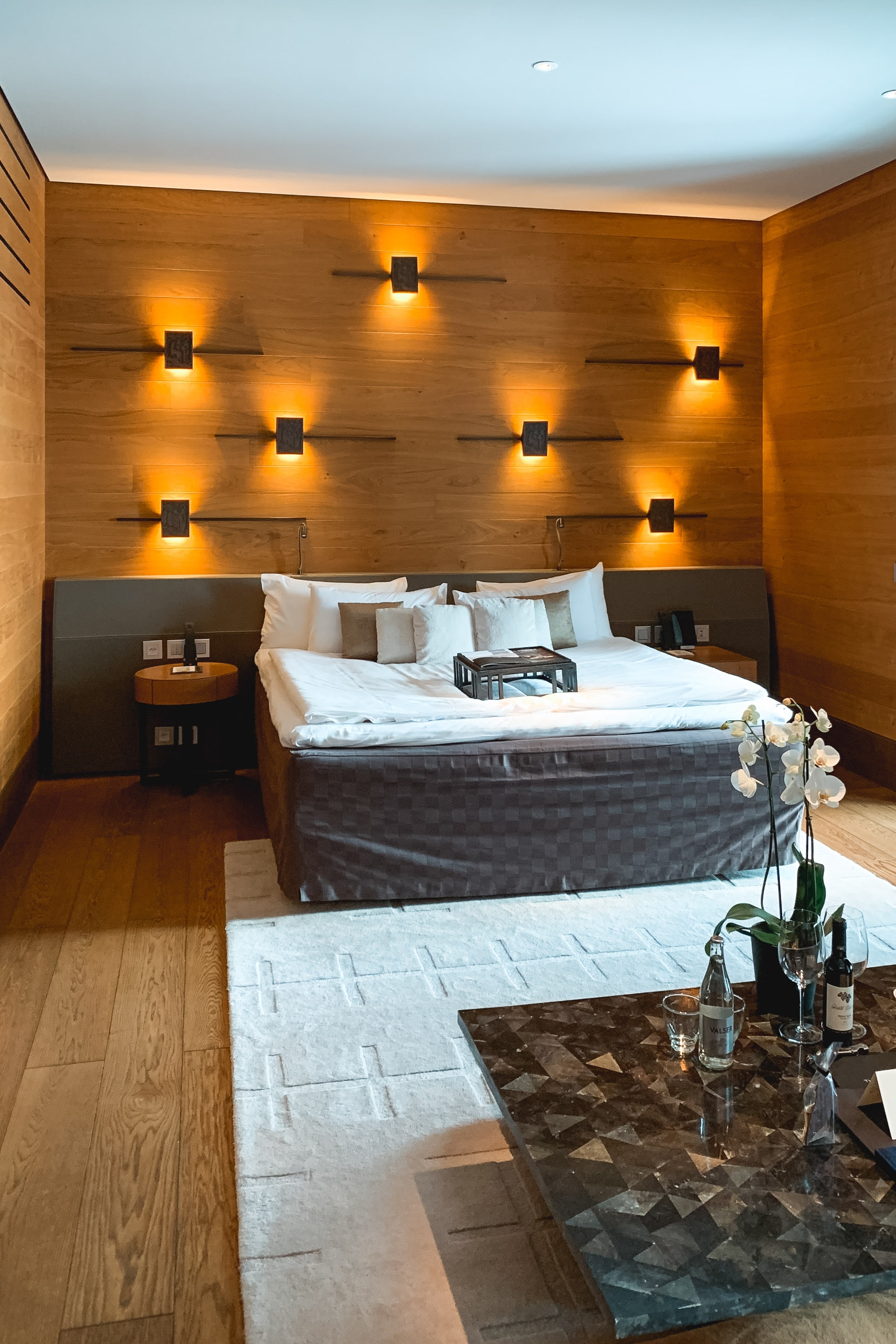 Modern bedroom with warm wood paneling, a centered platform bed with white linens, and symmetrically arranged uplighting creating an intimate amber glow against the feature wall