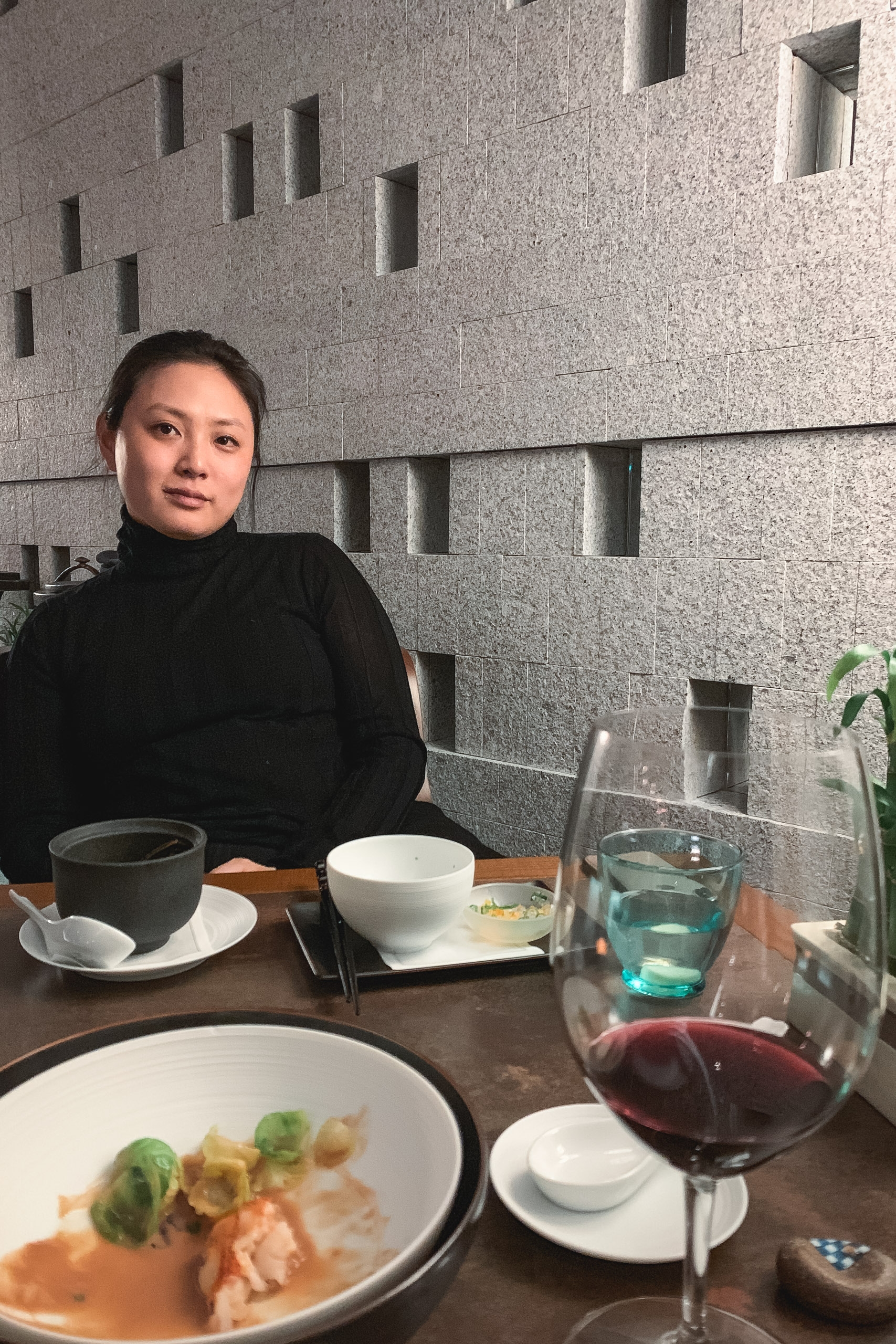 Woman in black turtleneck seated at dining table with plated food and wine glass, concrete wall with rectangular window openings behind her