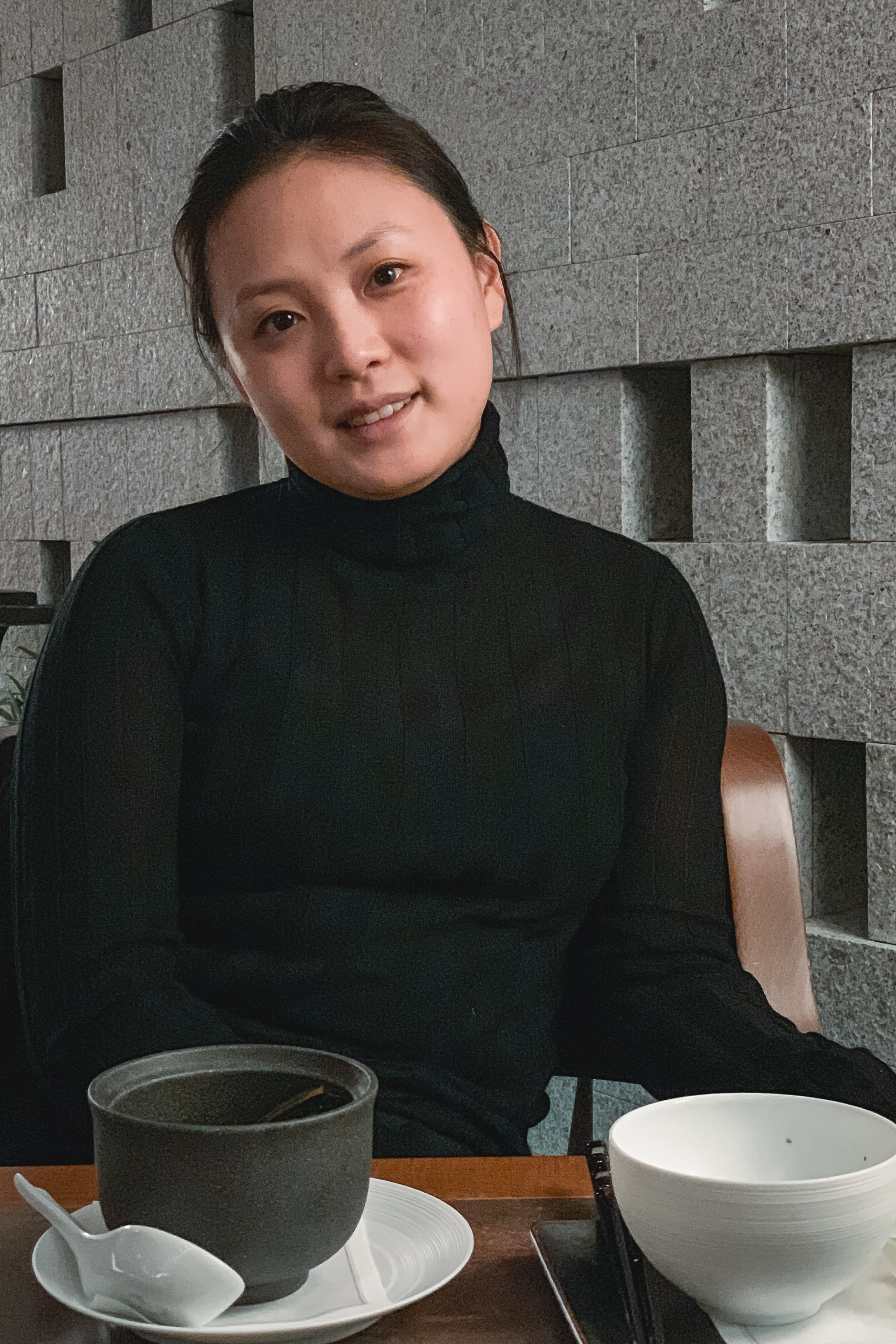 A woman in a black turtleneck sits at a wooden table with ceramic bowls and cups, smiling toward the camera against a geometric gray stone wall backdrop