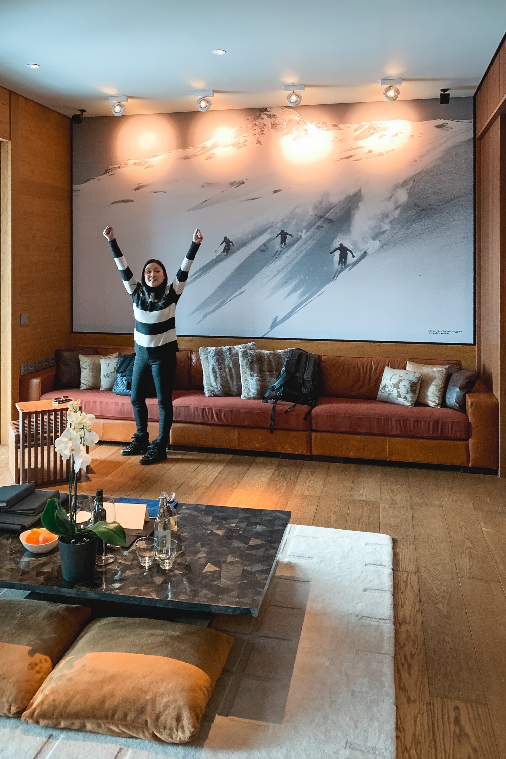 Woman celebrates with raised arms in a modern living room beneath a large mountain skiing mural, surrounded by warm wood paneling and an orange leather sofa.