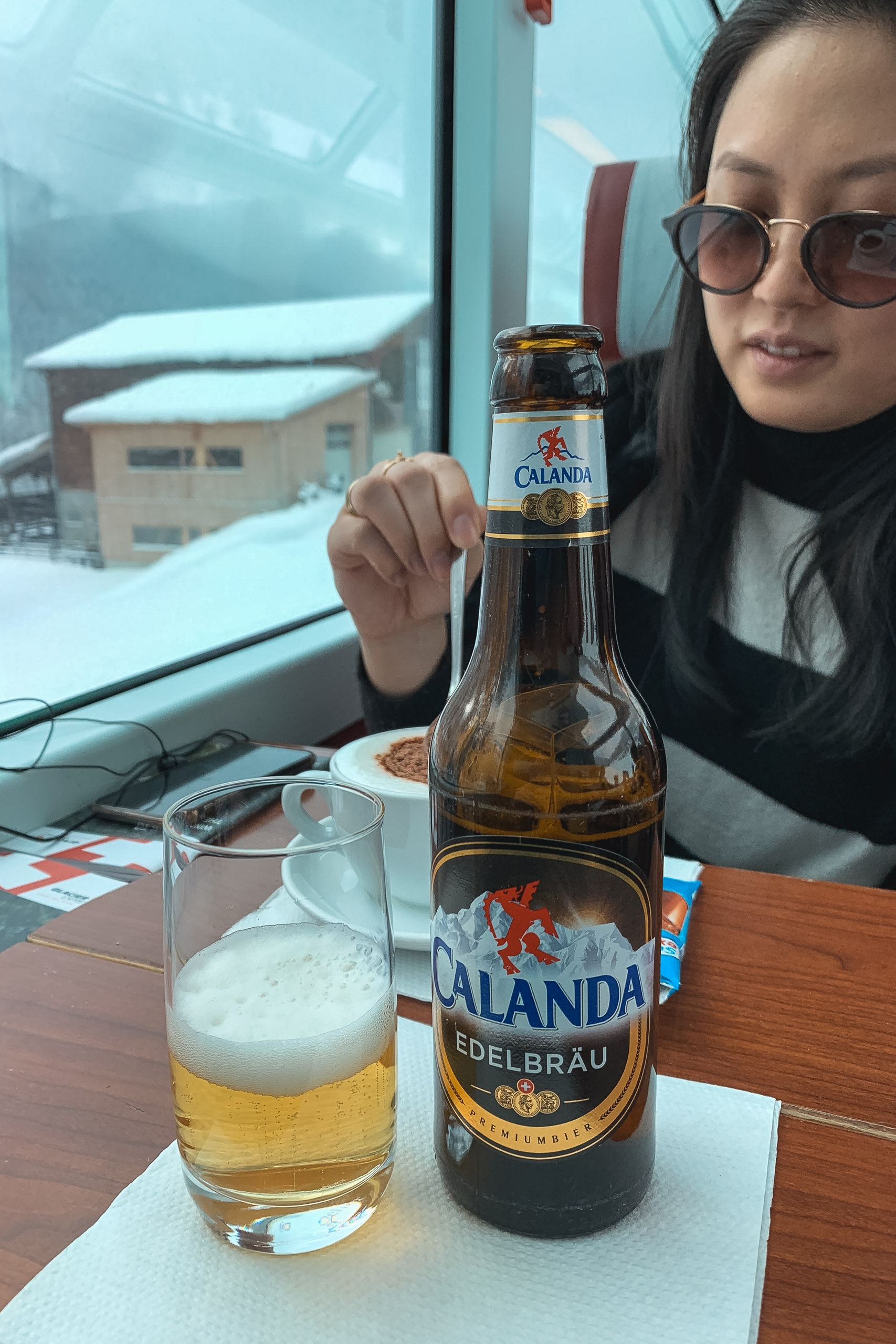 Woman wearing glasses holds a Calanda beer bottle at a table with snowy alpine landscape visible through large windows behind her