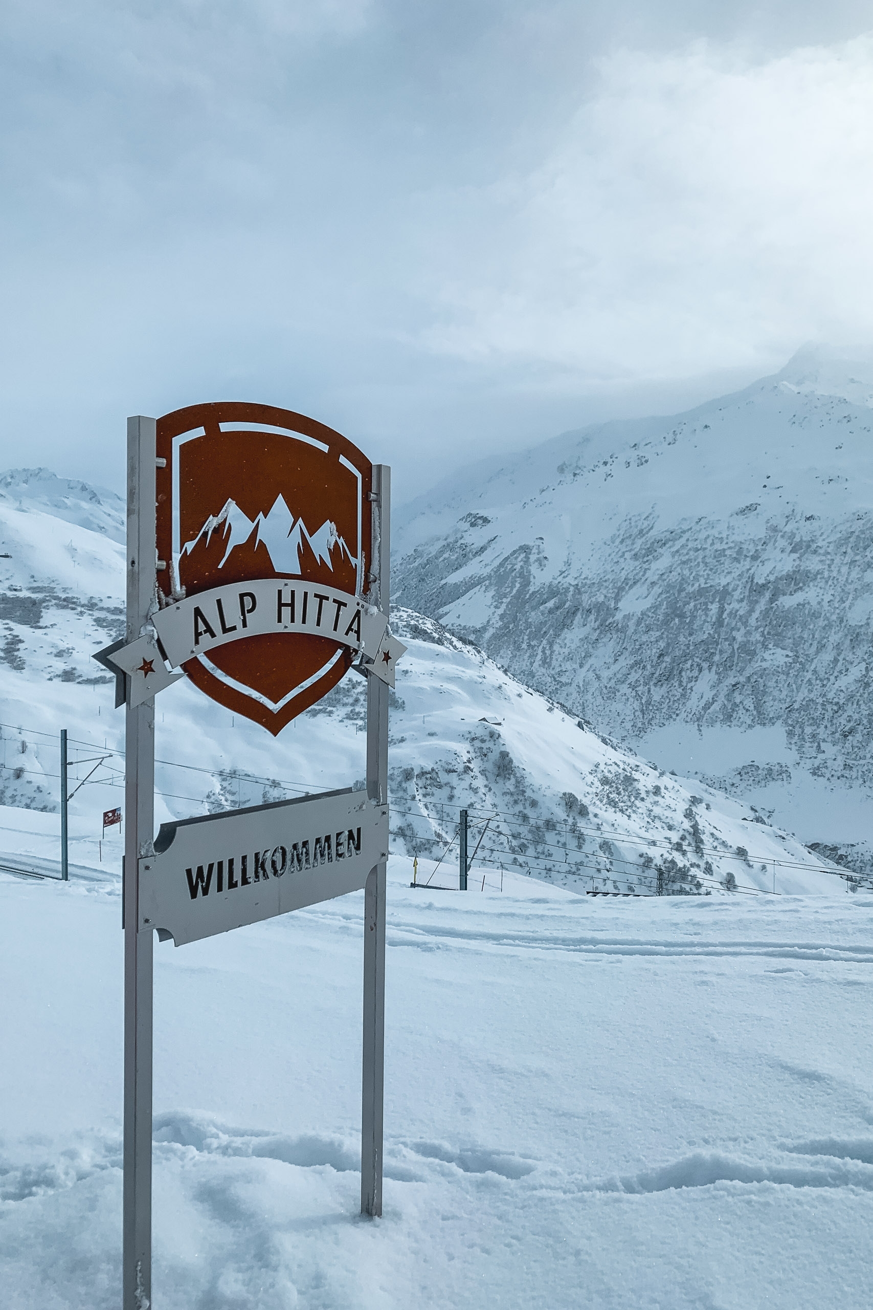 A wooden "Alp Hitta" welcome sign stands in snow-covered alpine terrain, with snow-laden mountains rising dramatically in the misty background under overcast skies