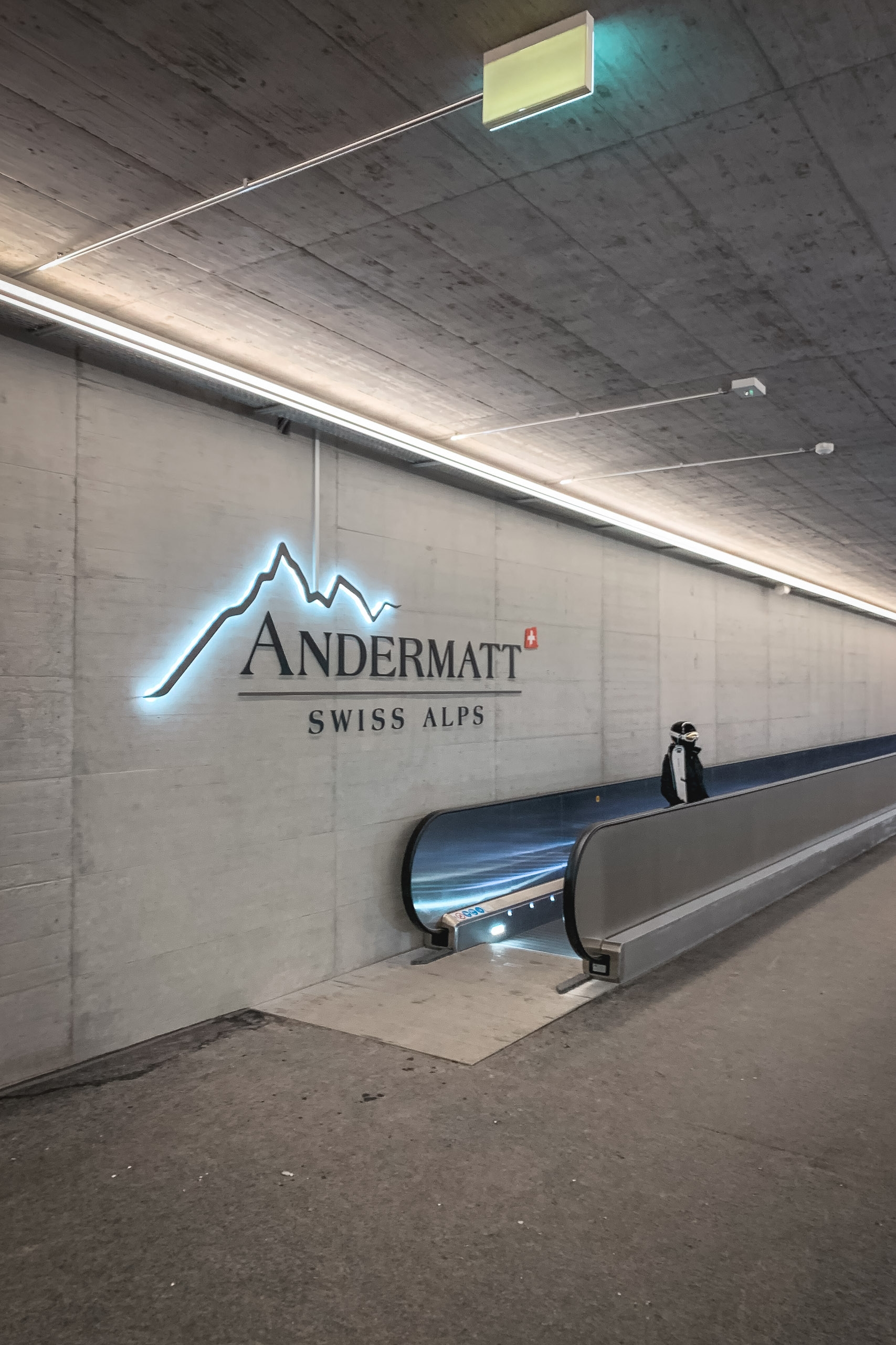 A modern underground tunnel features a blue neon-outlined mountain logo with "Andermatt Swiss Alps" text on a concrete wall, with an escalator and illuminated ceiling panels creating a contemporary transit aesthetic