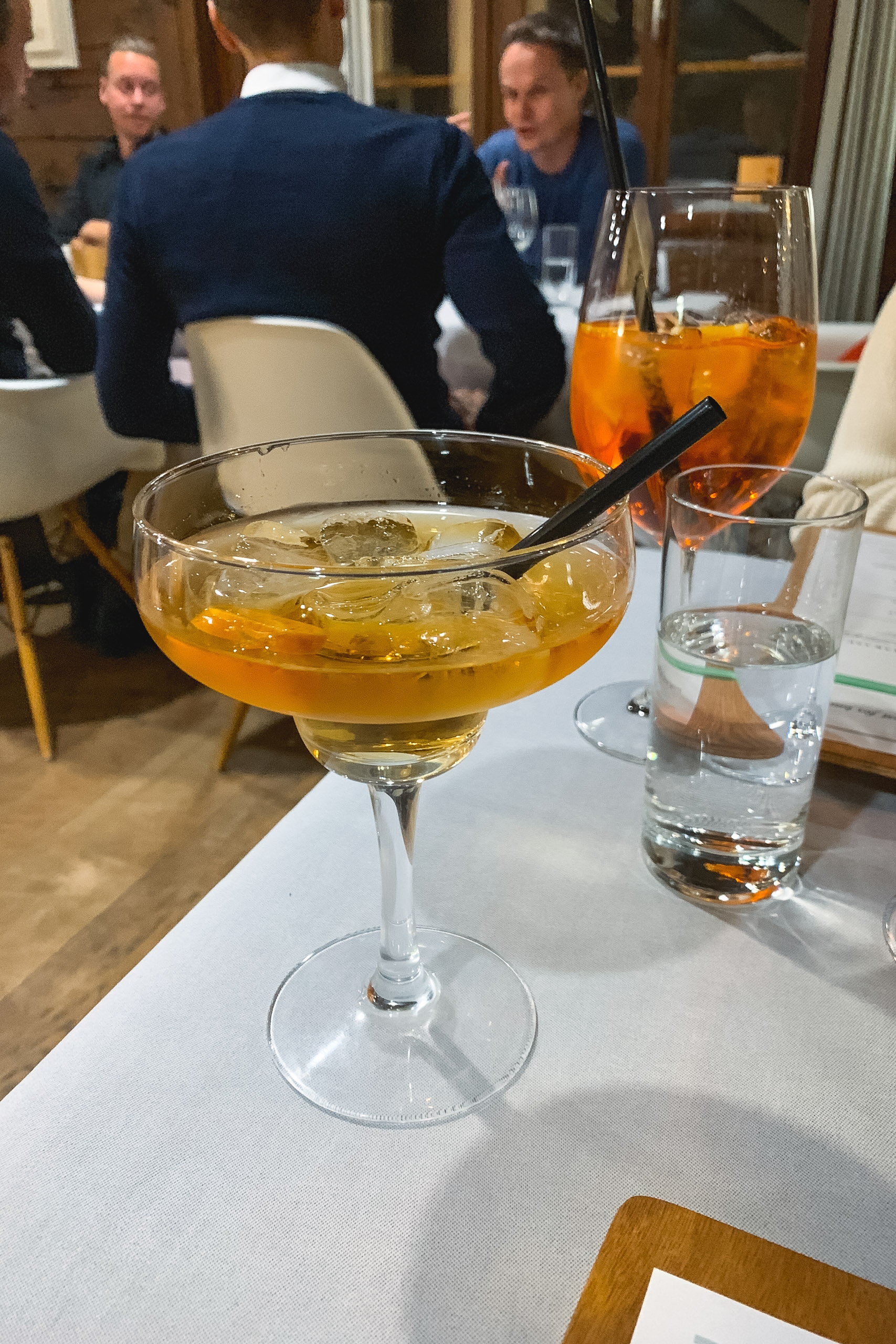 A golden cocktail in a coupe glass with a black straw sits on a white table, with blurred figures in dark sweaters conversing in the background of an indoor venue