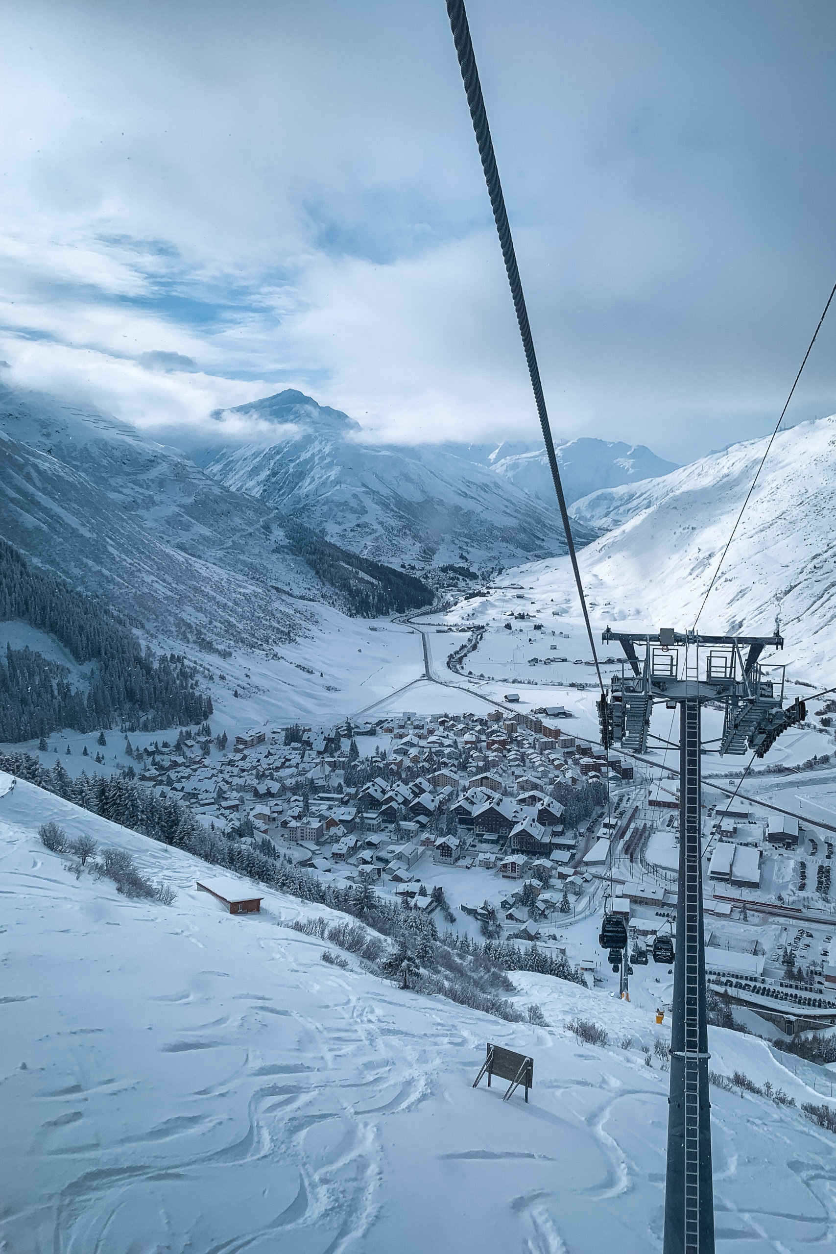 A ski lift cable stretches diagonally across snowy alpine terrain, with a snow-covered mountain village nestled in the valley below and snow-capped peaks rising in the misty distance