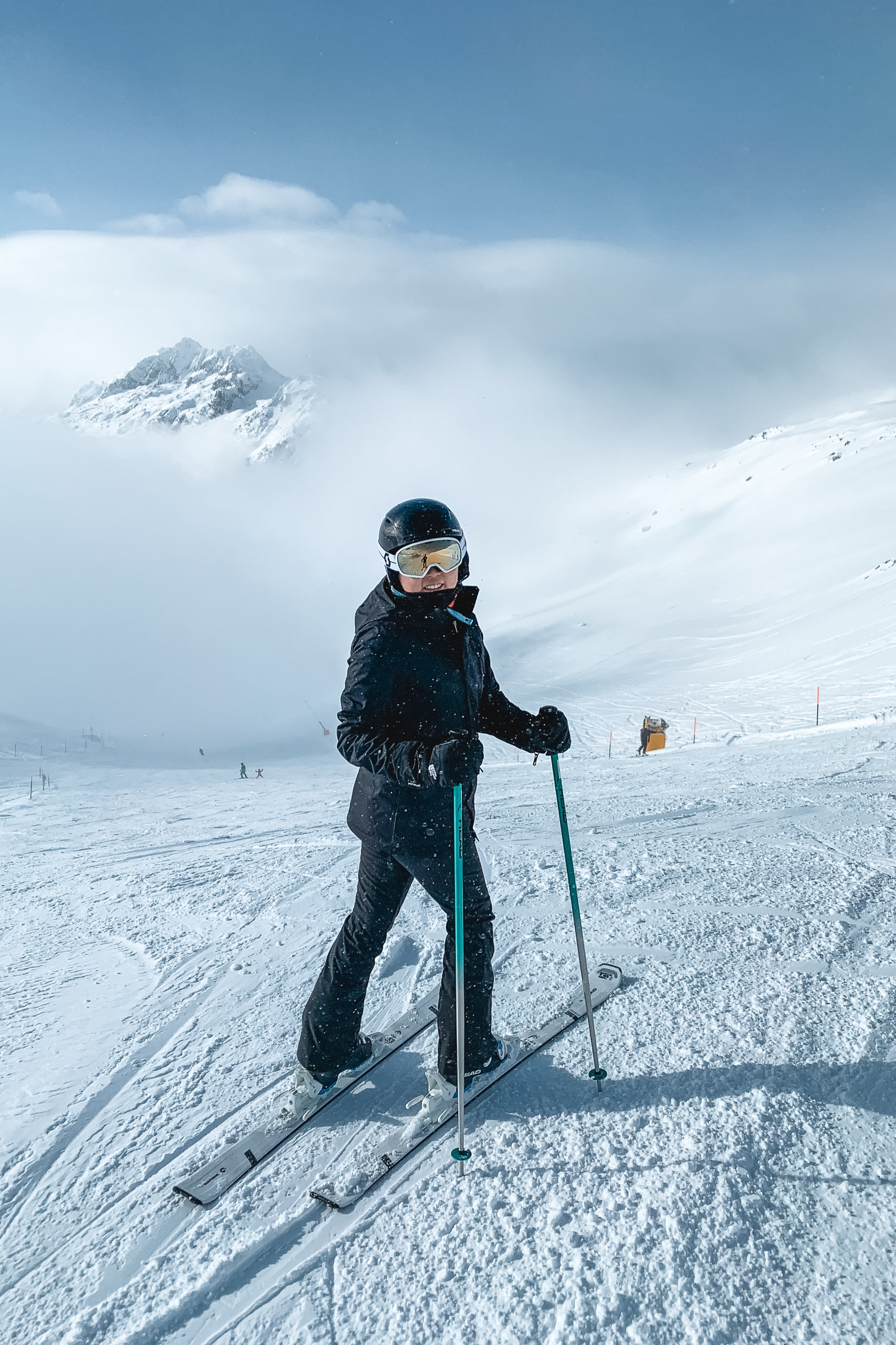 A skier in black gear stands on a snowy alpine slope facing the camera, with a snow-capped mountain peak emerging through clouds in the misty background under clear blue sky