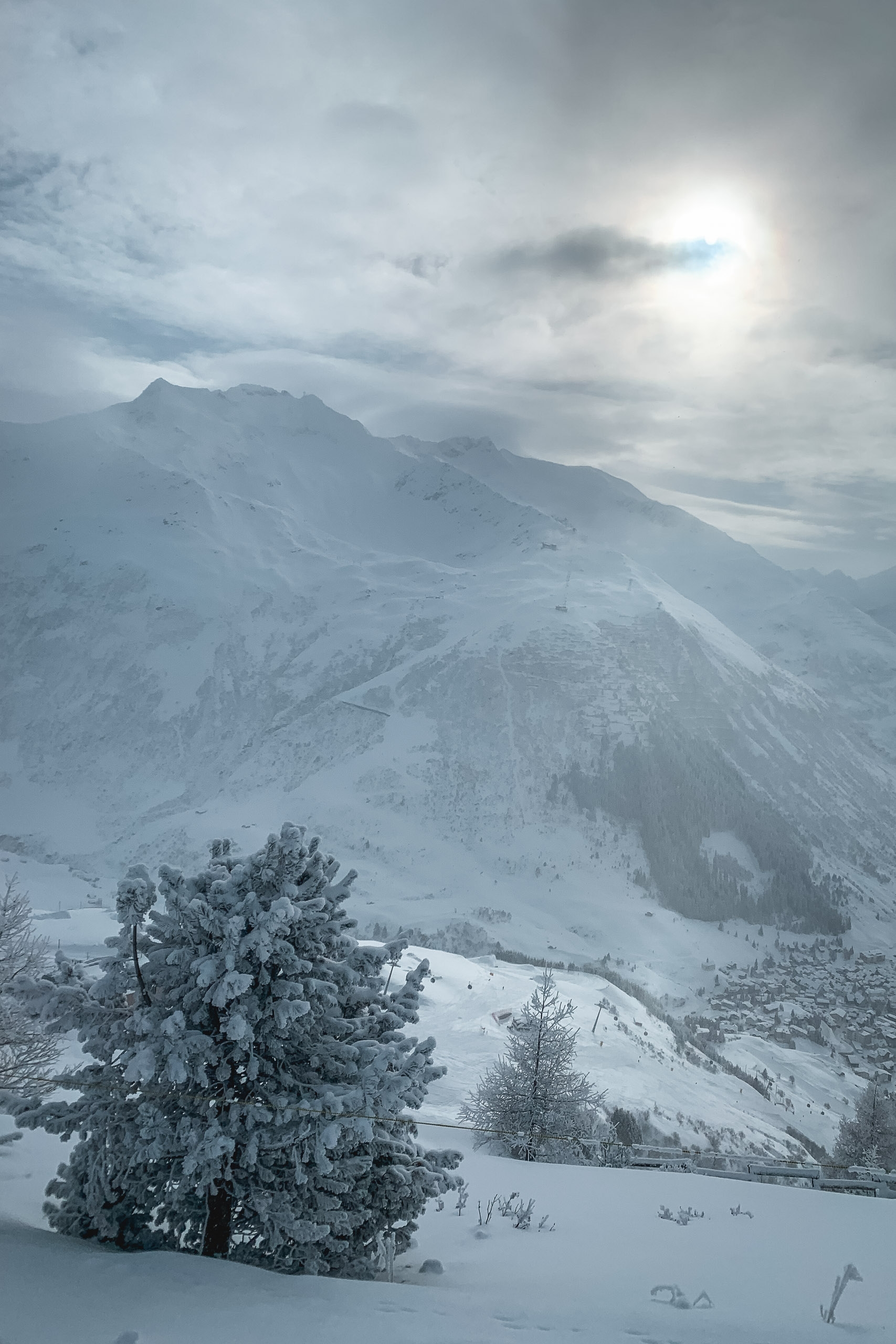Snow-laden evergreen trees dot a pristine white valley beneath a towering, snow-covered mountain under overcast winter skies with diffused sunlight breaking through clouds