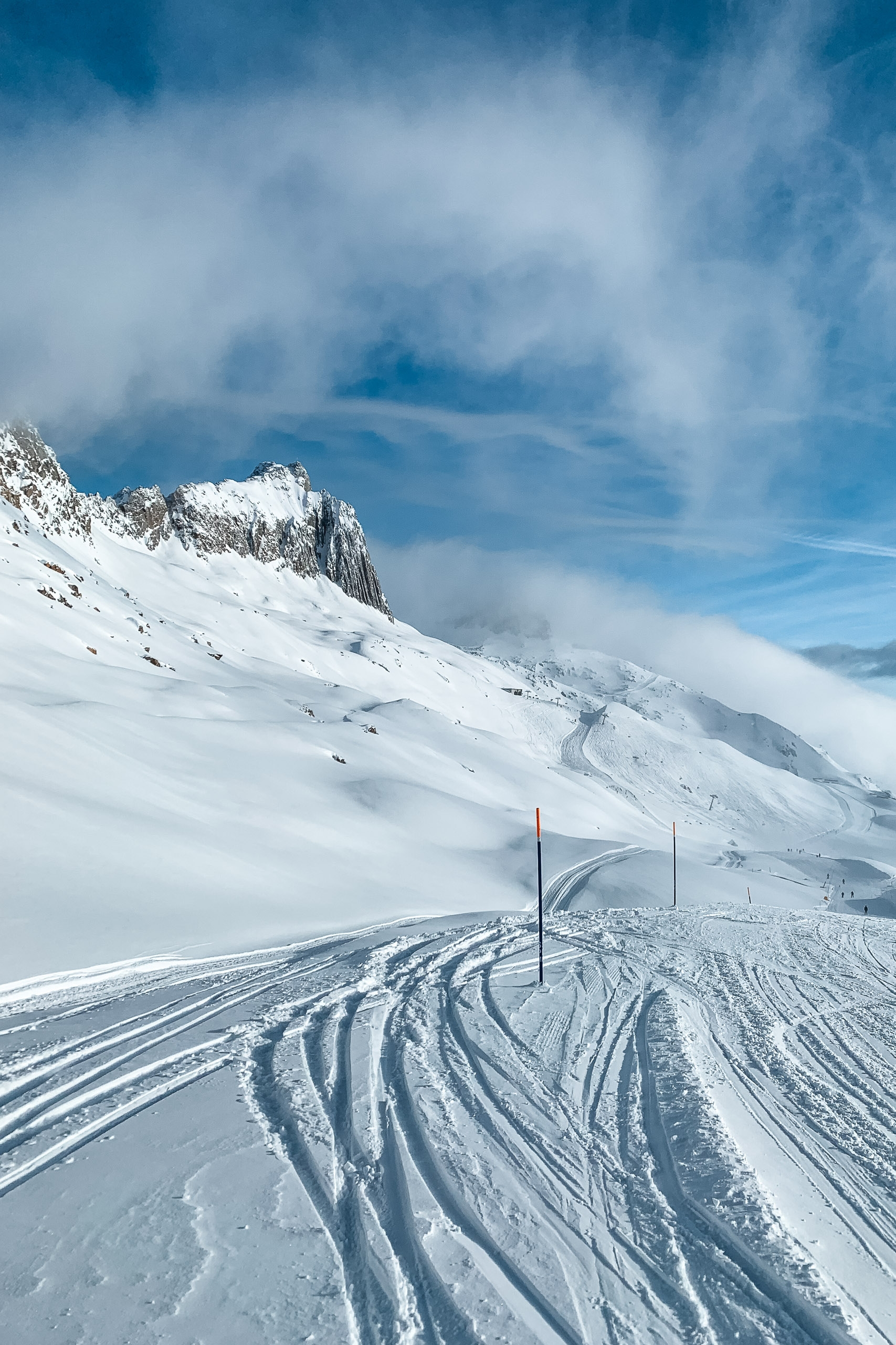 A snow-covered alpine slope descends toward a marked ski run bordered by red poles, with steep rocky peaks and rolling clouds filling the bright blue sky above