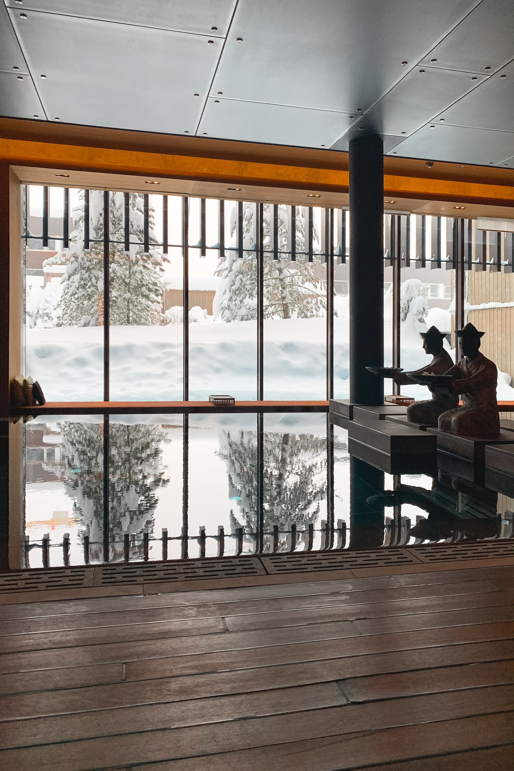 Two people sit on a bench beside expansive windows overlooking a snow-covered alpine landscape, with wooden flooring and modern ceiling panels framing the bright winter scene outside