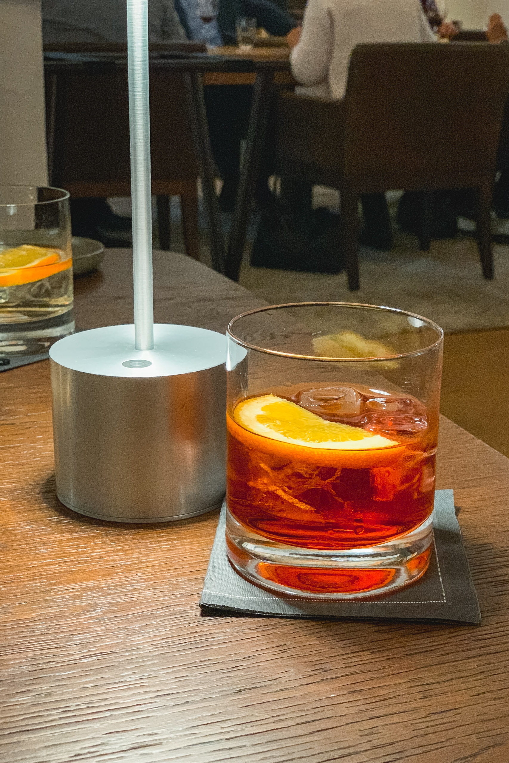 A glass of dark cola with an orange slice sits on a coaster beside a metallic cup with a straw on a wooden table in an indoor dining space