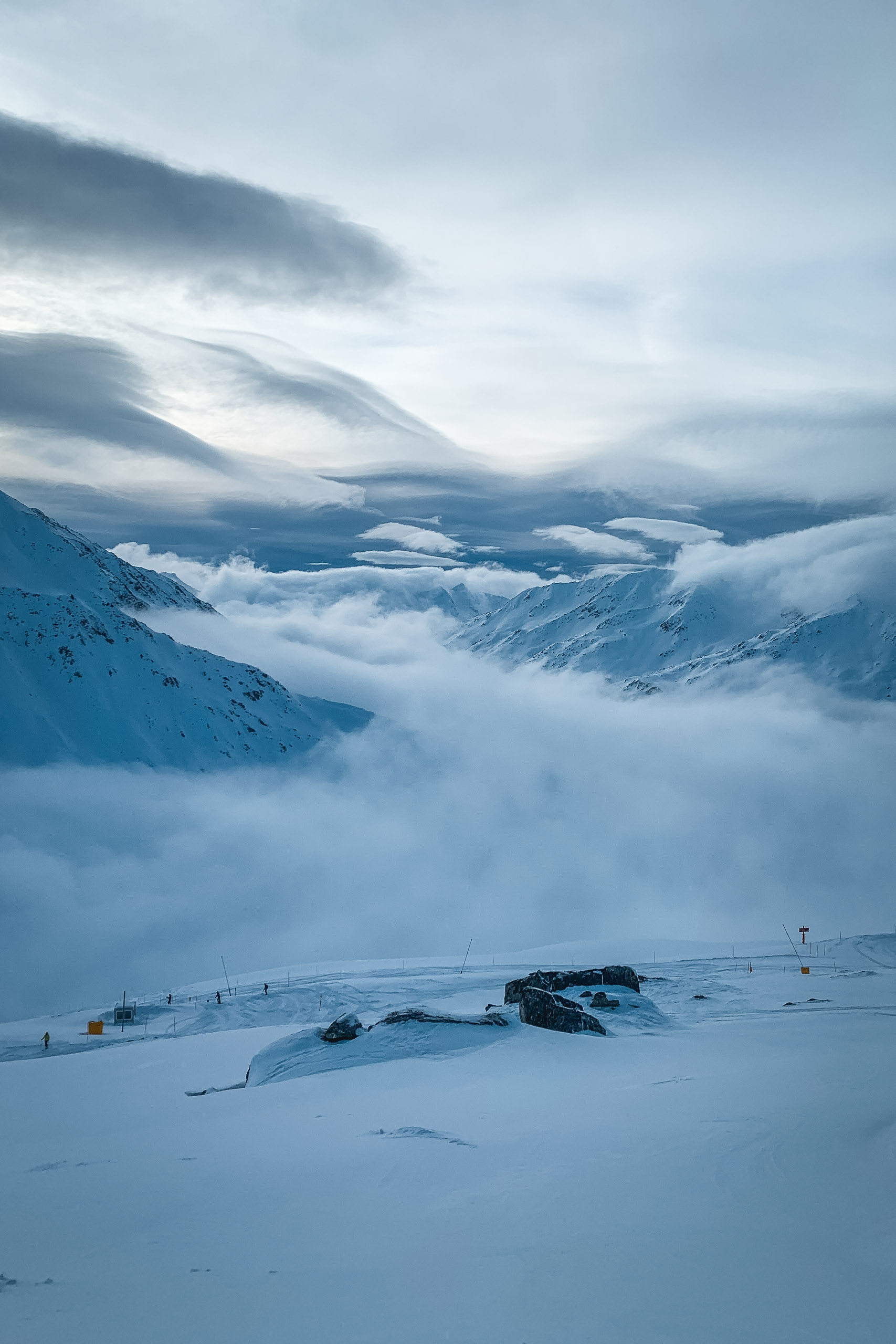 Snow-covered alpine plateau with exposed rocks and distant peaks shrouded in thick cloud formations under overcast sky