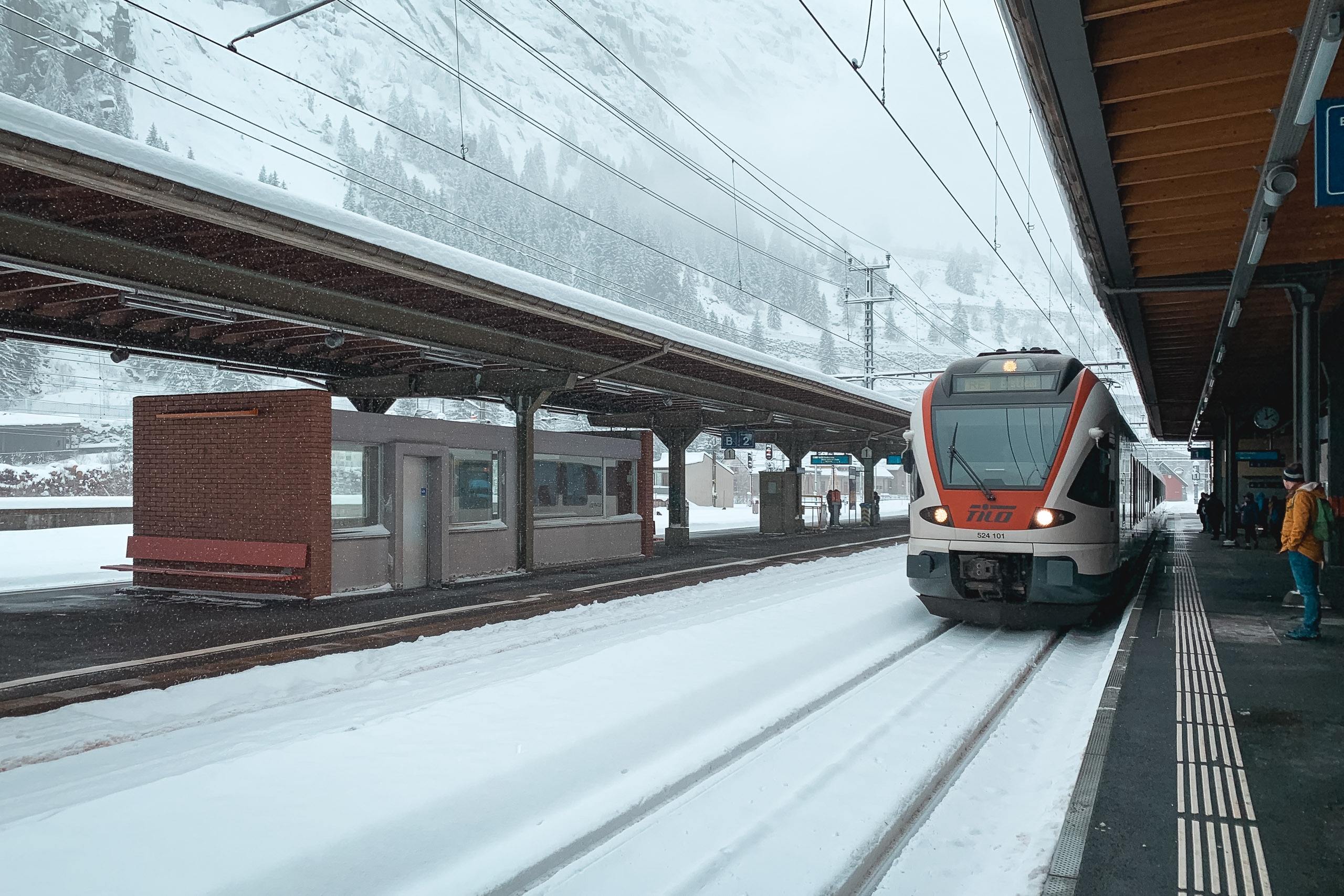 A red and white Swiss train sits at a snow-covered platform beneath a metal canopy, with forested mountains visible in the misty background and a brick station building to the left