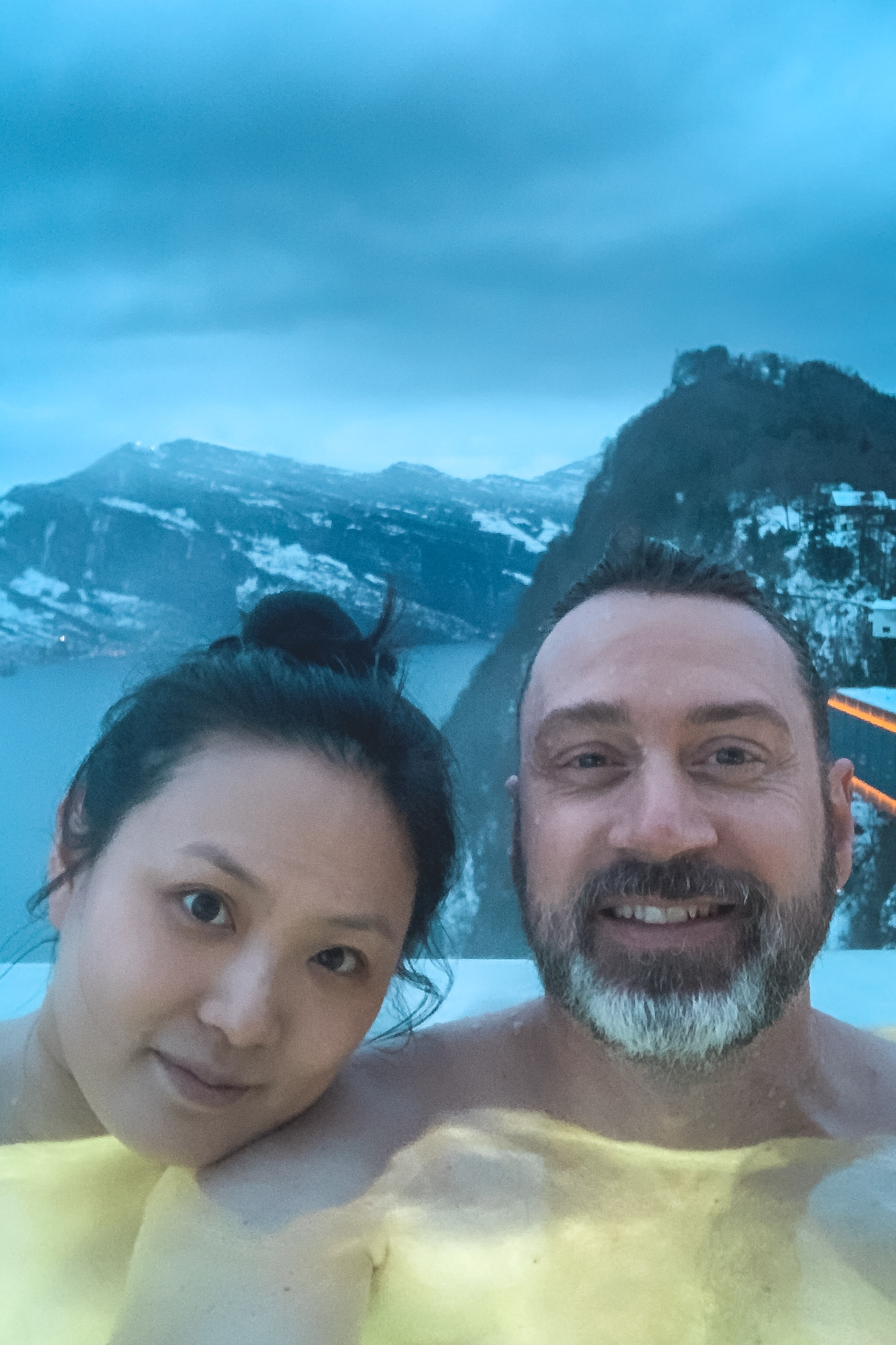 Two people smile at the camera while soaking in a hot spring with snow-covered Alpine mountains and a pristine lake visible in the misty background