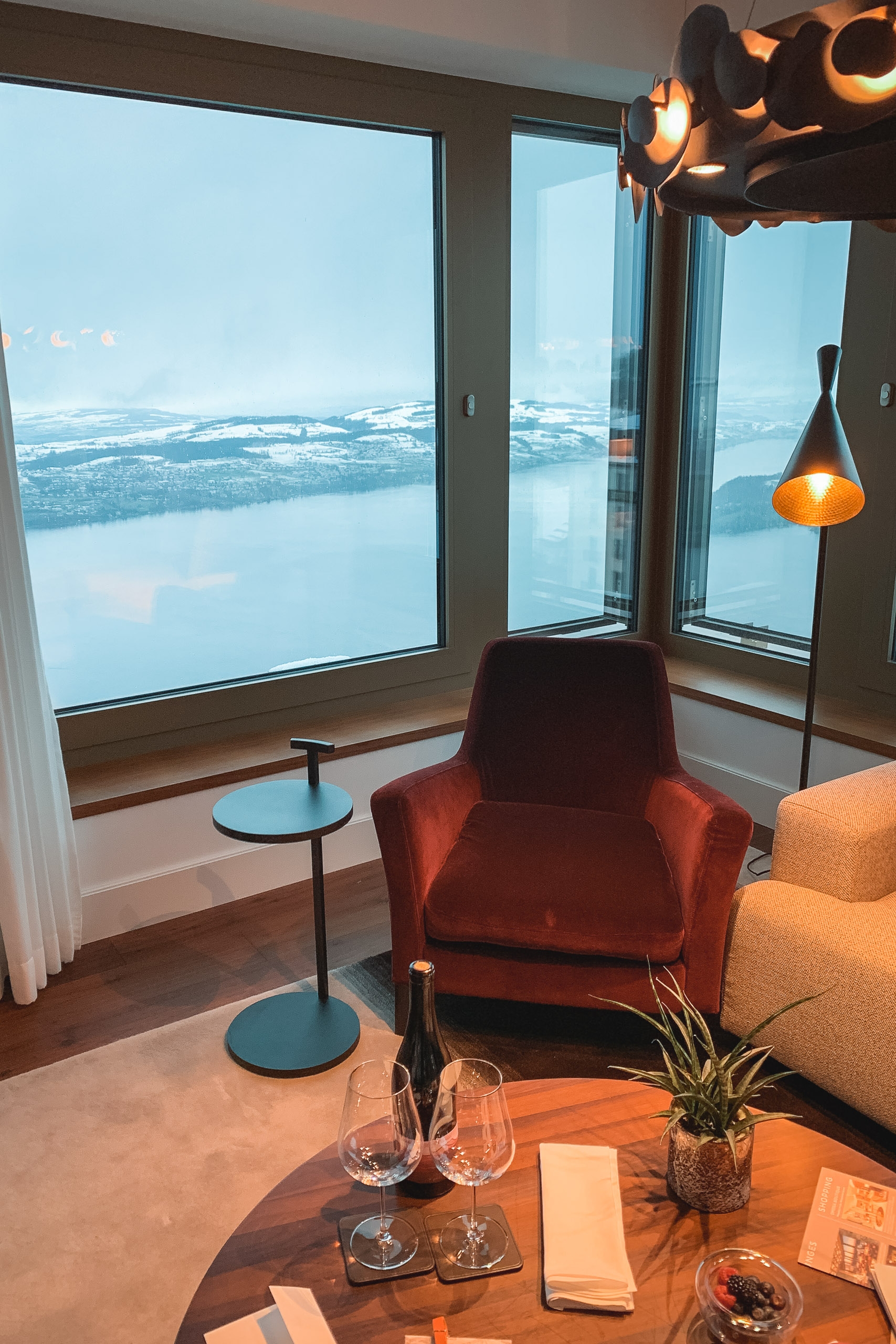 A modern interior room with a red velvet armchair and side table overlooks expansive alpine lakes and snow-capped mountains through floor-to-ceiling windows, with warm pendant lighting and contemporary furnishings below