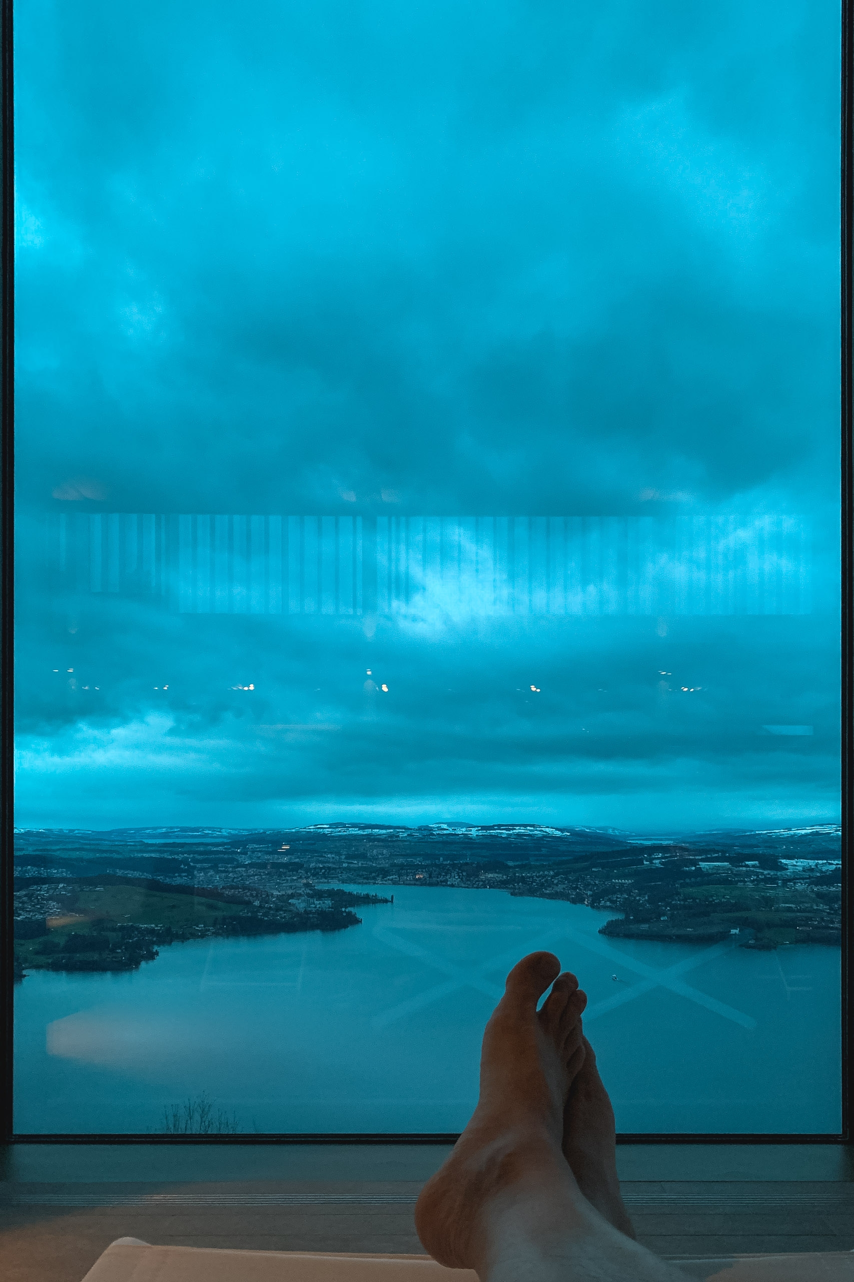 A person's bare feet rest on a wooden ledge while gazing through a large window at a turquoise alpine lake surrounded by forested shores and distant mountains under stormy blue skies