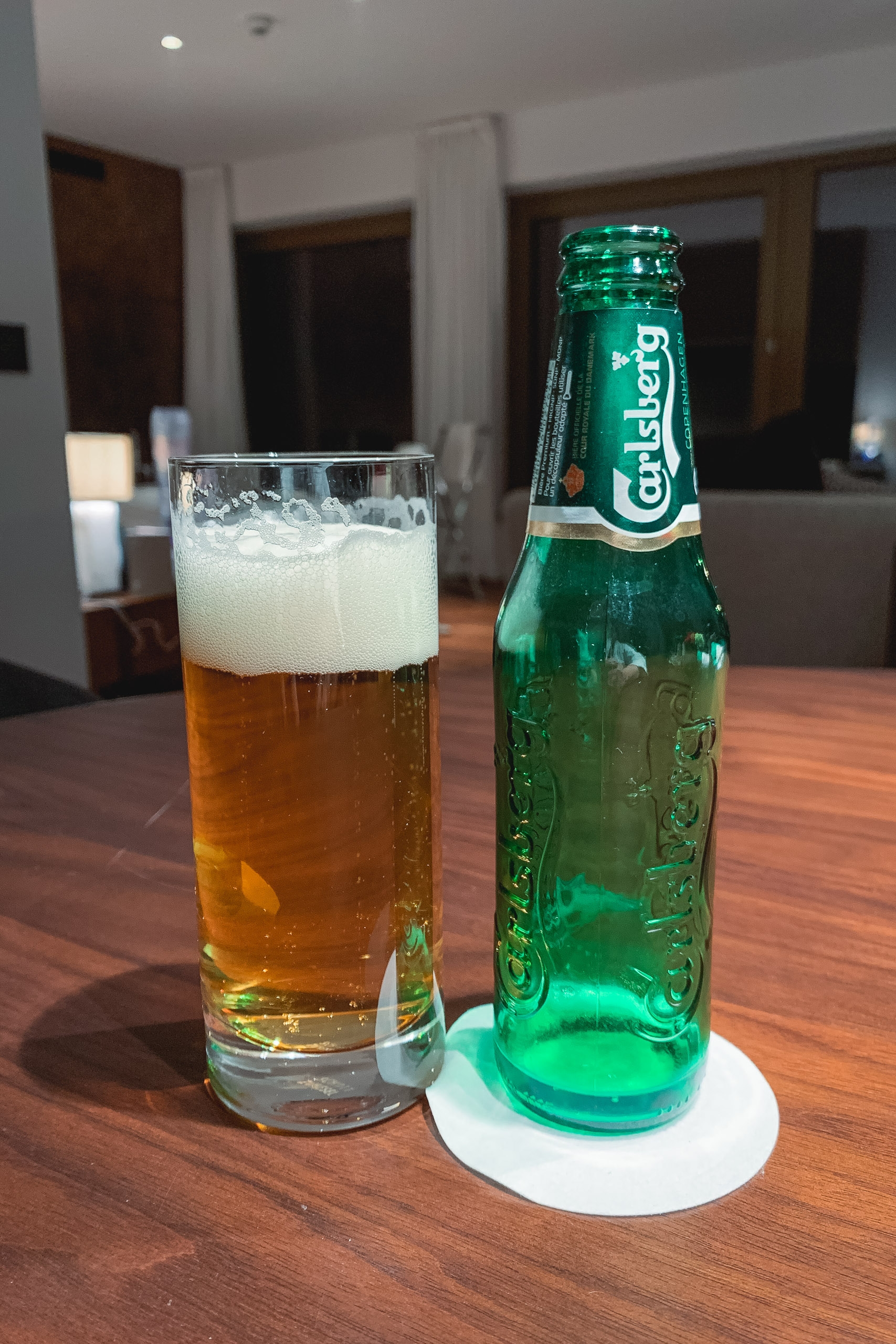 A Carlsberg beer bottle and freshly poured glass sit on a wooden table in a modern interior, with soft daylight filtering through windows behind them