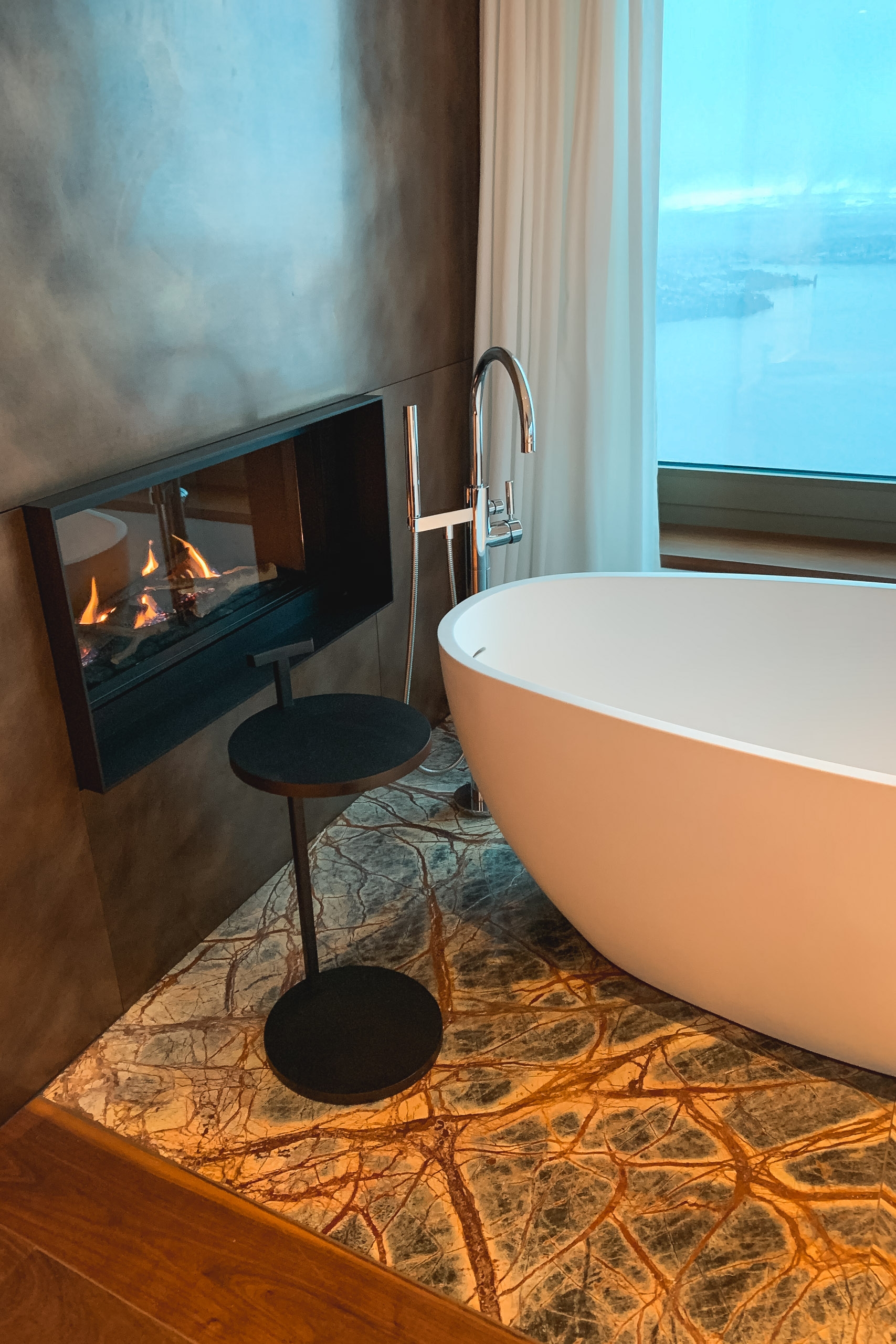 A modern luxury bathroom features a freestanding oval bathtub with chrome fixtures beside a contemporary fireplace, overlooking expansive blue lake and mountain views through floor-to-ceiling windows