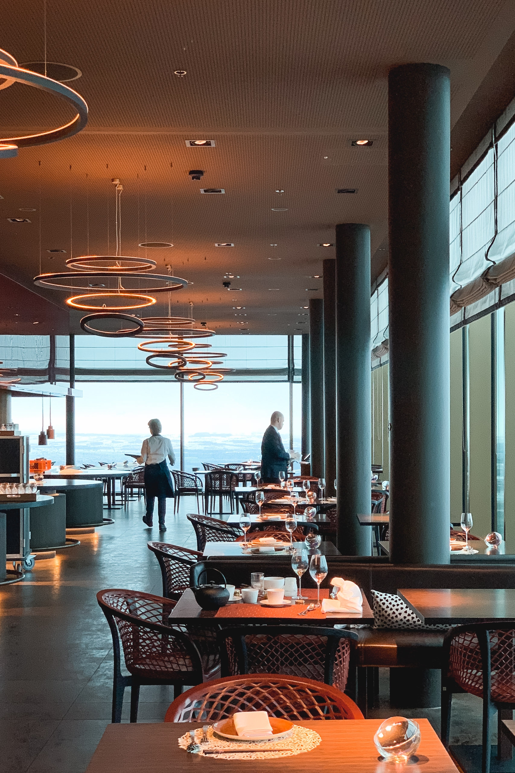 Modern restaurant interior with geometric pendant lighting, dark columns, and floor-to-ceiling windows overlooking a lake, featuring minimalist place settings and contemporary furnishings